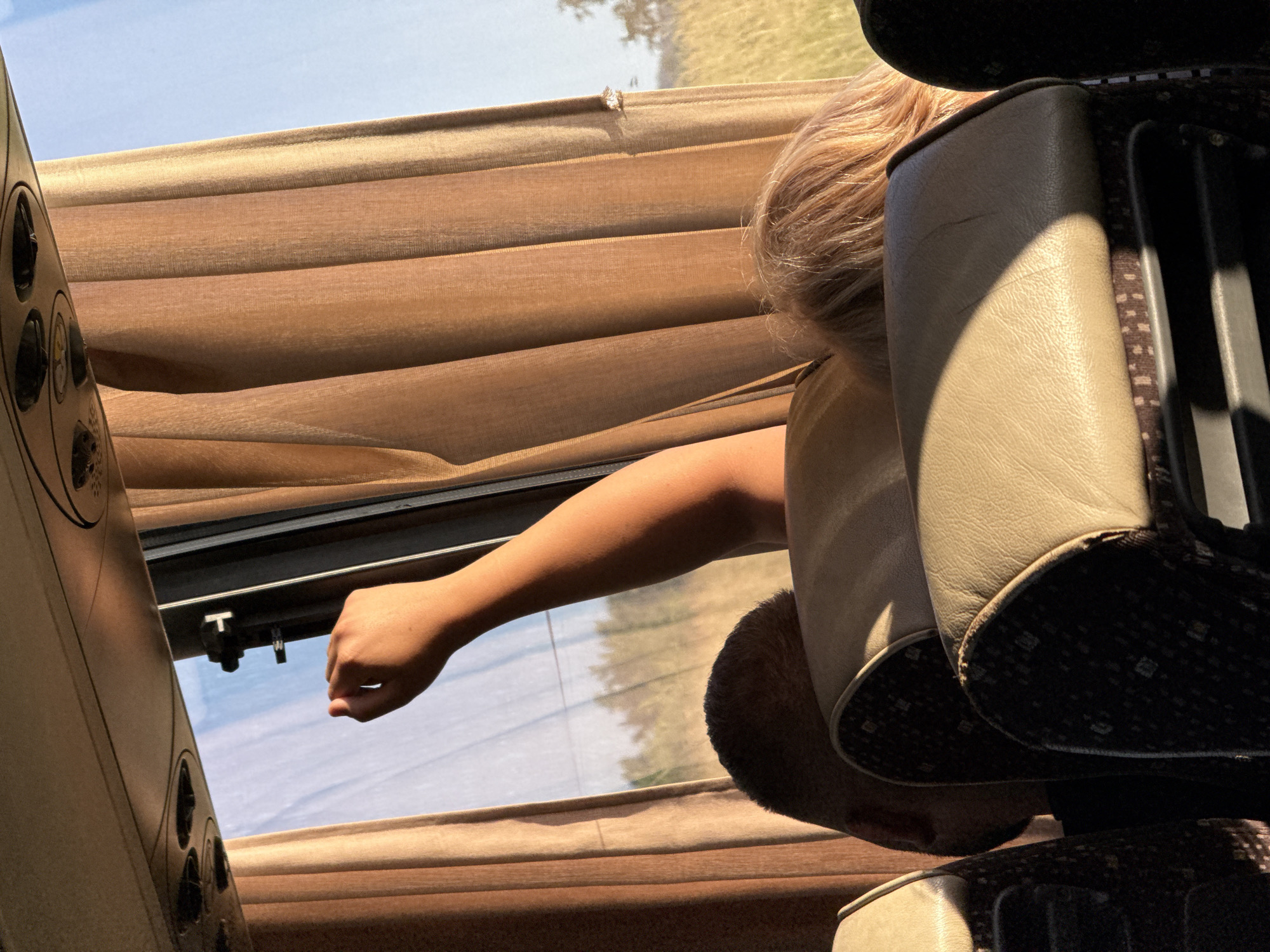 The image captures a scene from inside a bus, with a focus on the window area. A person's arm is extended out of the bus, with their hand near the top of the window frame. Brown curtains are drawn to the side, revealing a sunny sky and a field of grass and trees in the distance. The interior of the bus shows seats, with the headrests of two seats visible in the foreground. The lighting suggests it is either early morning or late afternoon, with warm sunlight casting shadows. The exact location of Kiosevtsi, Bulgaria, cannot be determined solely from this image.