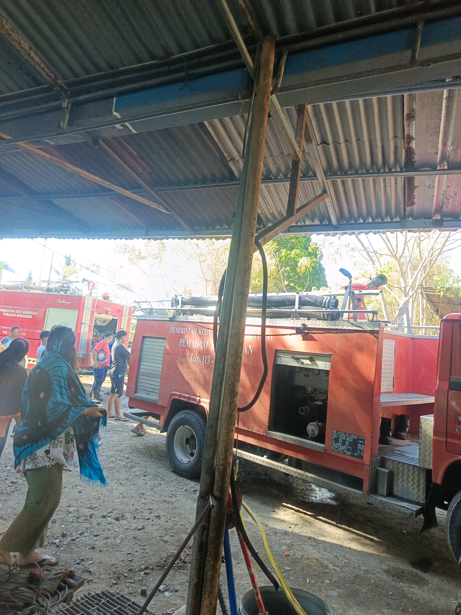 The image depicts a scene in what appears to be a fire station or depot in Gorontalo, Indonesia. Two red fire trucks, identified with text indicating "PEMERINTAH KOTA GORONTALO PEMADAM KEBAKARAN" (Gorontalo City Government Fire Department) and "PEMERINTAH KODY" (City Government), are parked under a corrugated metal roof supported by metal poles. One fire truck in the foreground has a nozzle mounted on a tripod on its roof and appears to be operational. The second fire truck, partially visible behind the first, also displays "PEMERINTAH KOTA GORONTALO PEMADAM KEBAKARAN".
Several individuals are present. In the foreground, a person wearing a patterned blue and black scarf over a white and floral patterned top and olive green pants is facing away from the camera, holding a blue scarf. To the left, partially obscured, are other individuals, one wearing a dark hooded garment and another in a dark top and light-colored pants. In the middle ground, between the two fire trucks, several men are standing. One man in a blue t-shirt and dark shorts is looking towards the fire trucks. Another man in a dark grey t-shirt and black shorts is also present.
The ground is a mixture of dirt and gravel, with scattered debris. There are various hoses and equipment visible on the ground, including a bucket with yellow and red hoses. The environment appears to be outdoors, with trees visible in the background under a bright sky. The overall impression is a functional but somewhat unkempt operational area for the fire department.
