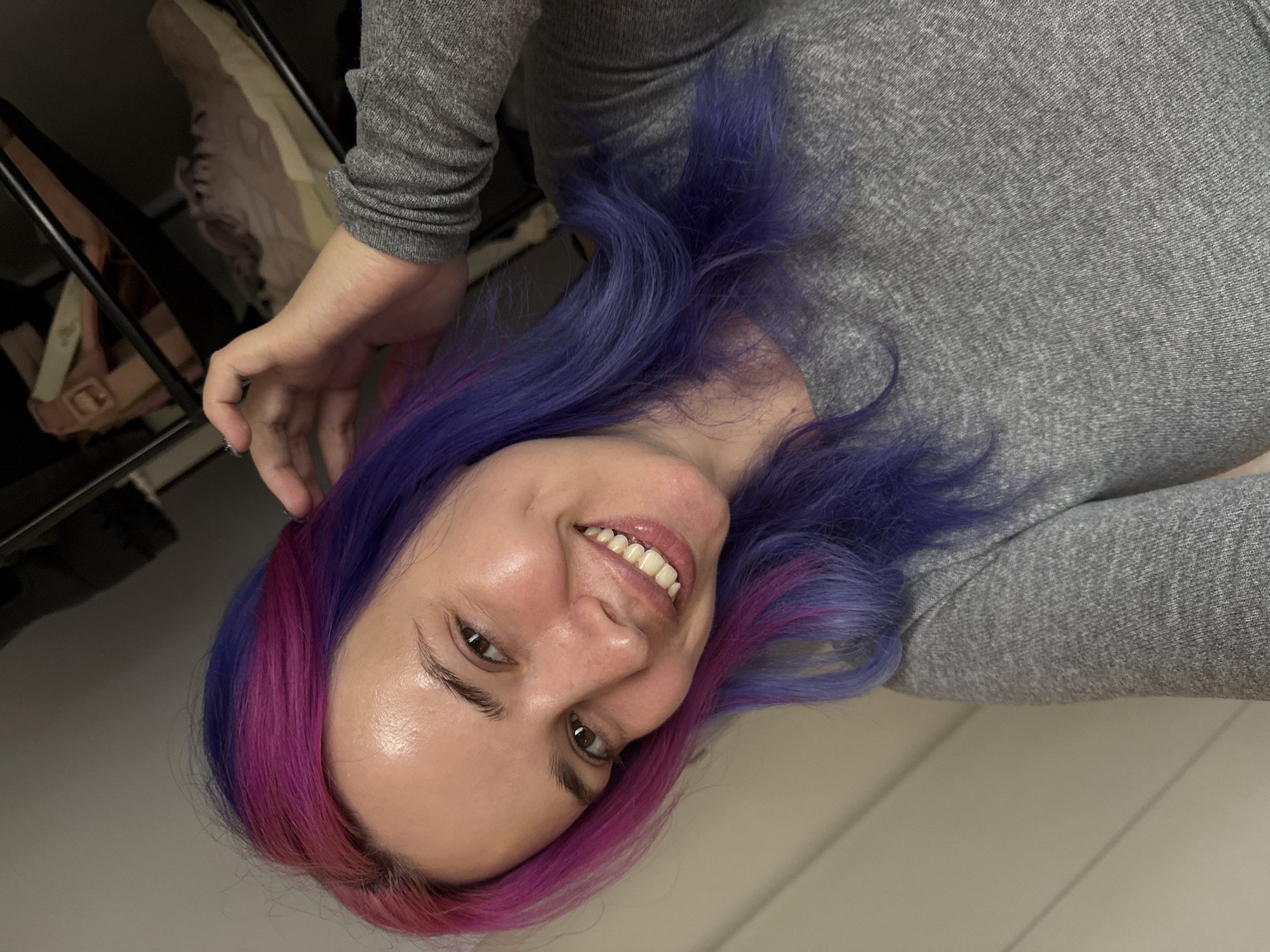 An eye-level portrait shows a woman smiling broadly towards the camera, her head tilted slightly. She has vibrant bi-colored hair, transitioning from purple to bright pink or magenta, which frames her face and shoulders. She is wearing a grey, long-sleeved top with a visible ribbed texture. Her face appears bright with a dewy complexion, and her brown eyes are open and looking forward. Her wide smile reveals her teeth, conveying a joyful and friendly emotion. One hand is partially visible near her head, with a ring on one of her fingers.
The background is divided into two main sections. To the left of the frame, there is a dark, somewhat cluttered area that appears to be an open closet or shelving unit, with glimpses of light-colored items, possibly shoes, stored within. To the right, a plain, light-colored wall or surface is visible. The scene is set indoors, likely within a residence in the City of Zagreb, Croatia. No specific time of day or weather conditions are discernible, and there is no visible text.