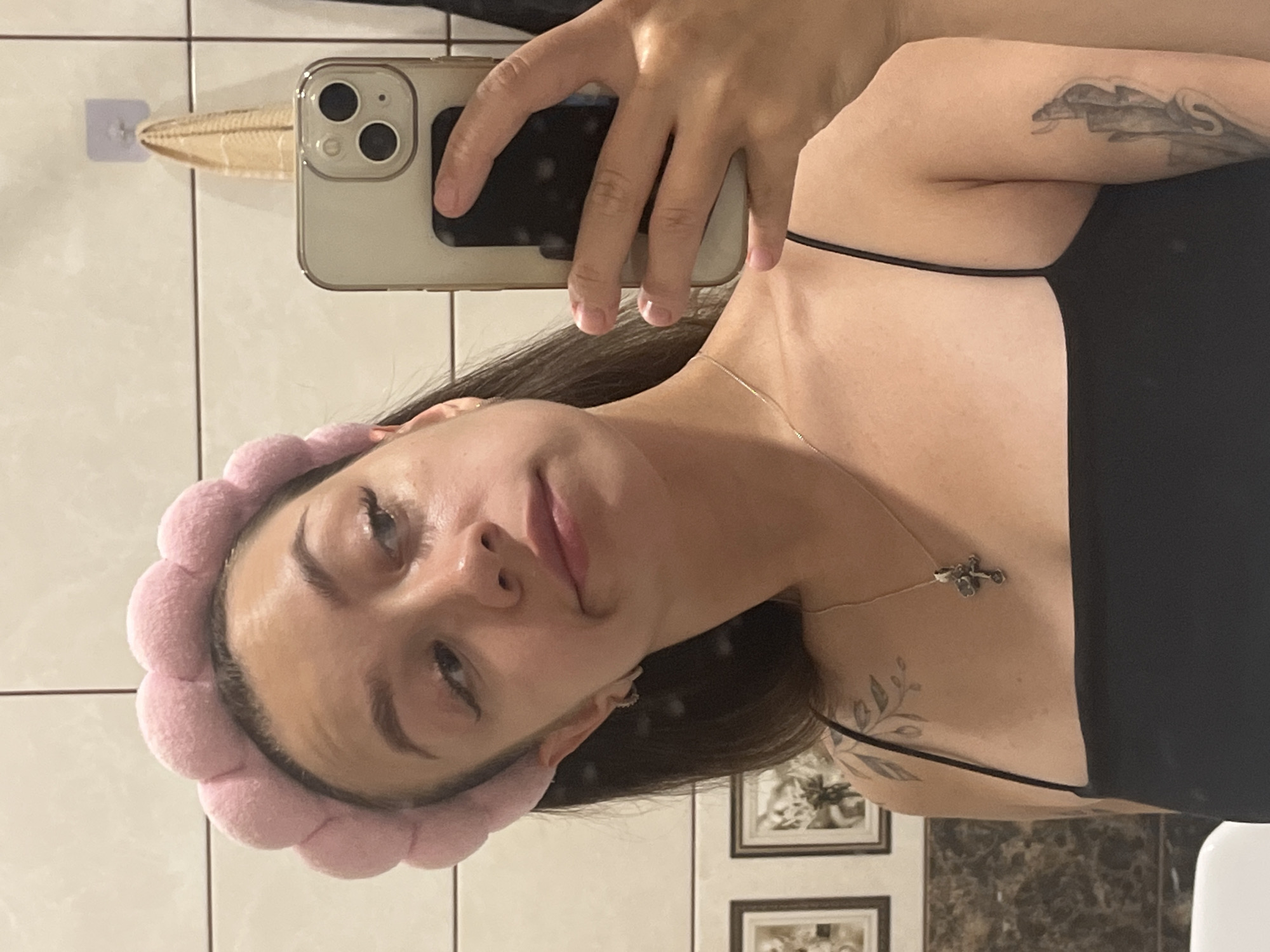 A young woman, wearing a pink fluffy headband and a black spaghetti strap top, takes a selfie in a bathroom mirror. She has dark hair, light skin, and appears to be looking away from the camera. A silver necklace with a cross pendant is visible around her neck. She has tattoos on her left shoulder and right arm. The bathroom features white tiled walls, a marble countertop, and framed pictures on the wall. A light-colored towel hangs from a hook.