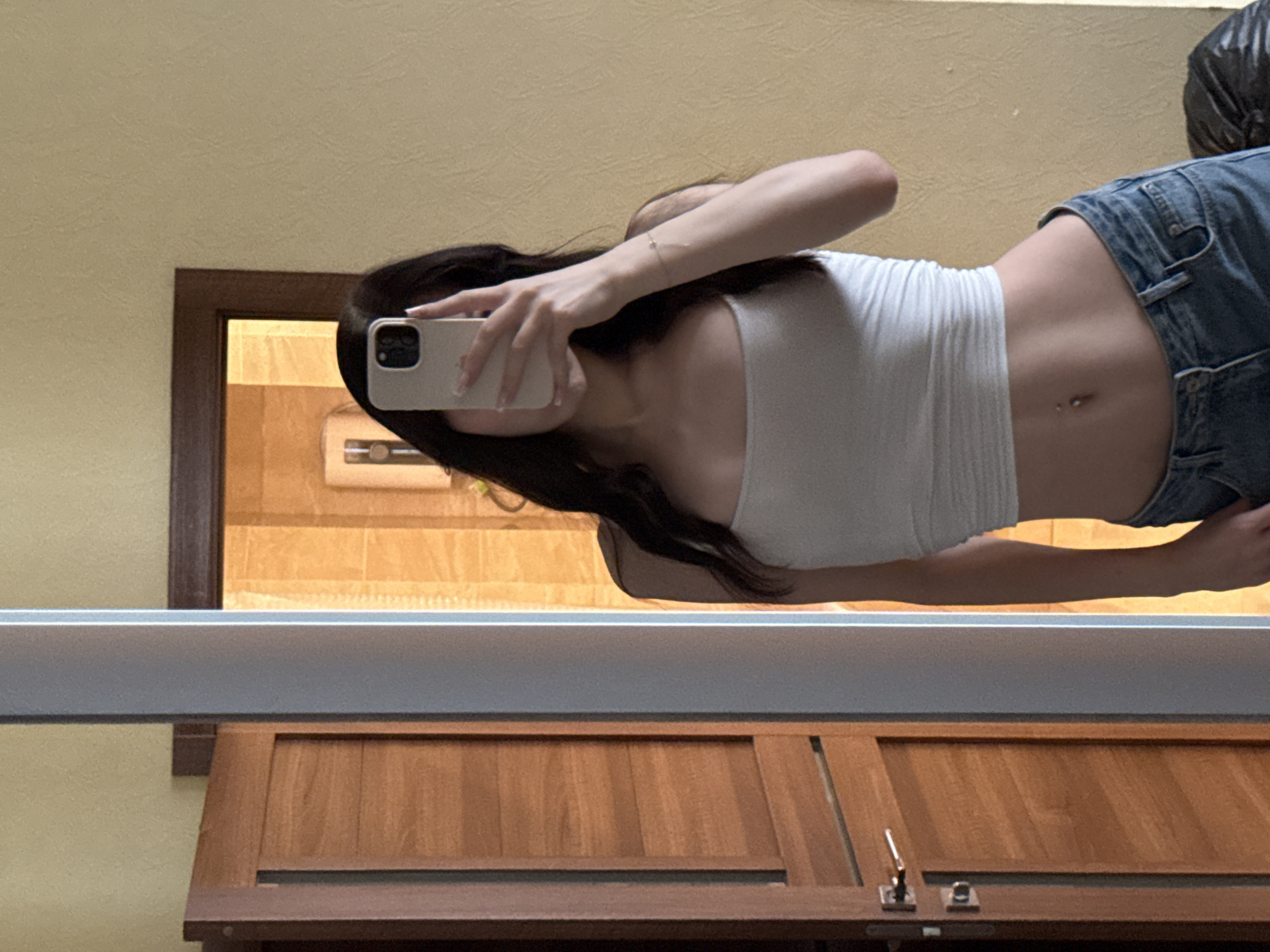 A young woman with long dark hair is taking a mirror selfie in a room that appears to be a bathroom or dressing area. She is wearing a white crop top and blue jeans. Her midriff is exposed, revealing a navel piercing. She is holding a smartphone in her right hand to take the photo. The room features a wooden door on the left and a mirrored wall on the right. The background reflected in the mirror shows tiled walls, suggesting a bathroom. The overall lighting is warm. This scene is in a residential apartment in Khimki, Russia.