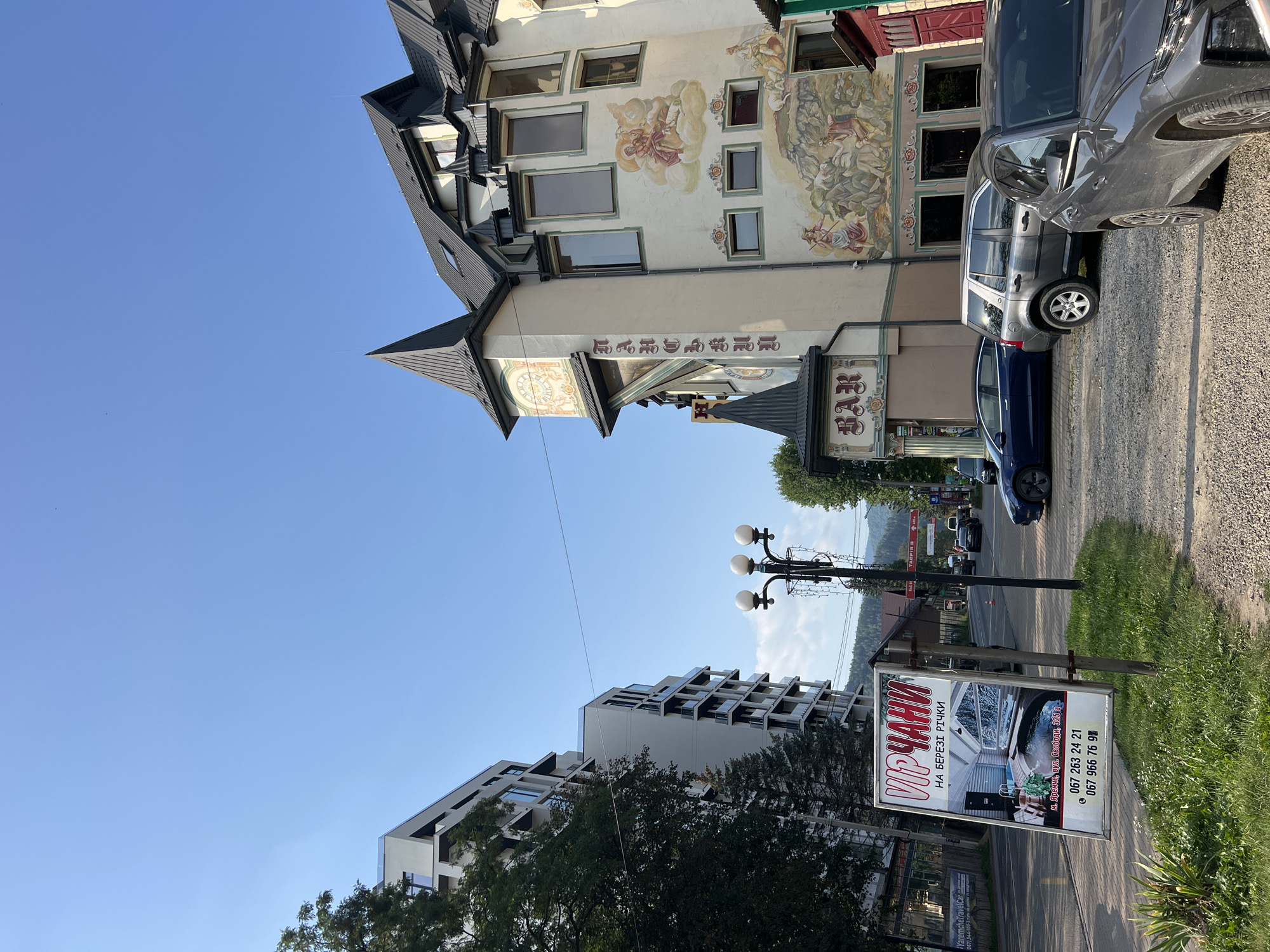 The image depicts a street scene in Yaremche, Ukraine, on a clear, sunny day. In the foreground, on the right, several cars are parked on a gravel lot, including a dark grey SUV and a dark grey crossover. A tall, white billboard with red lettering and images stands prominently on the left, advertising "VIP ЧАНИ" (VIP Chas) and "НА БЕРЕЗІ РІЧКИ" (On the River Bank), along with contact information and a picture of a hot tub.
Behind the billboard, a modern, multi-story apartment building with white facades and balconies rises on the left. To the right of this building, a distinctive, multi-story building with a peaked turret and intricate murals adorns its facade. The building features a clock on one side and a sign with the word "BAR" in decorative lettering. Murals depict figures in what appear to be historical or mythological scenes, with elements of nature. A street lamp with three globes stands between the billboard and the colorful building.
The street stretches into the background, with more buildings and trees lining its sides. A range of hills is visible in the distance. The sky is a bright, cloudless blue. The overall impression is a blend of modern and traditional architecture in a town setting, likely a tourist area given the prominent advertisement.
