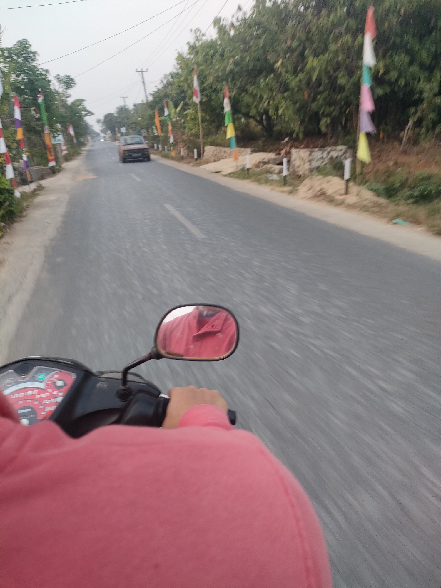 The image depicts a first-person perspective of riding a motorcycle on a road in Sukoyoso, Indonesia. The road is paved and appears to be a two-lane street with a white dividing line visible. On either side of the road, there are trees and foliage, with some small structures or walls. Numerous Indonesian flags, predominantly red and white, are planted along the roadside. In the distance, a light brown car is driving away from the viewer. The rider is wearing a pink long-sleeved shirt and is holding onto the handlebars of the motorcycle. A rearview mirror is attached to the handlebars, reflecting the rider's red shirt. The sky is overcast, suggesting a hazy or cloudy day. The motion blur indicates that the motorcycle is in motion.