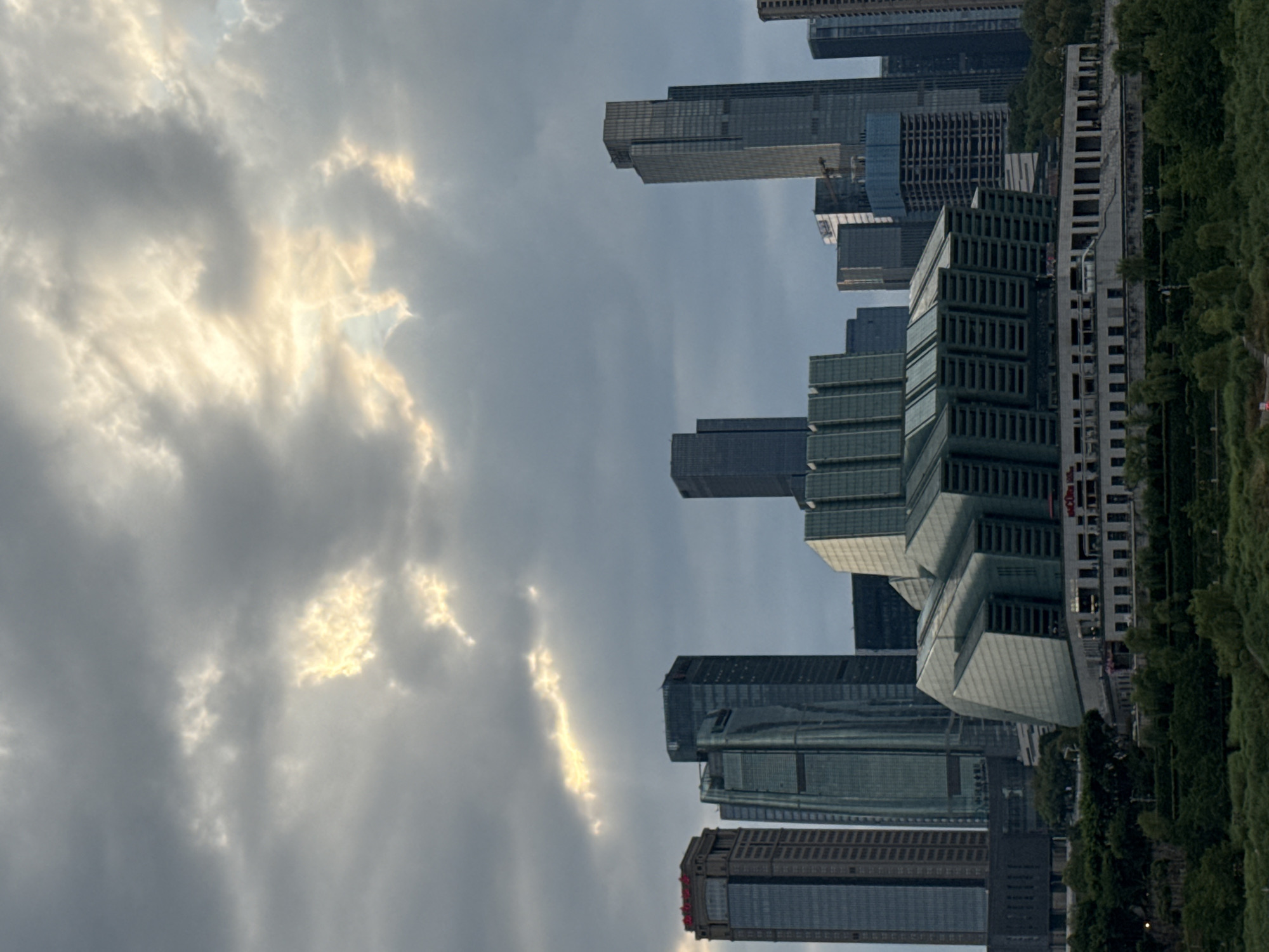 The image shows a city skyline with modern buildings and a cloudy sky. The buildings are of varying heights and architectural styles, with some appearing to be skyscrapers. The sky is filled with dramatic clouds, with rays of sunlight breaking through in some areas. The foreground of the image features a lush green landscape with trees and vegetation. In the distance, the city of Nan'an District, China, is visible, characterized by its contemporary architecture and urban development. The scene captures the grandeur of the city and the dynamic nature of the sky.