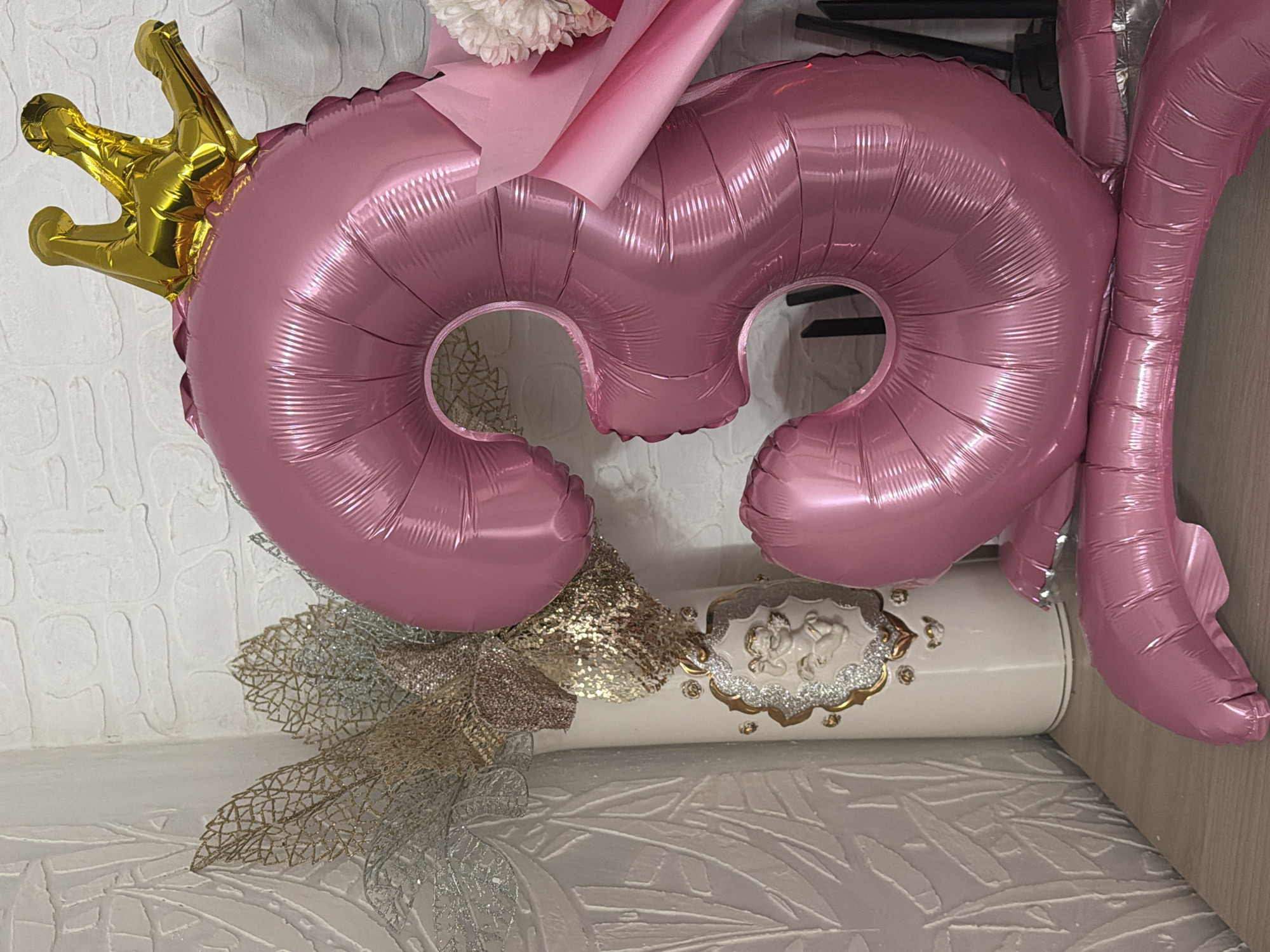 The image shows a pink number 3 balloon with a golden crown on top, next to a pink number 6 balloon. A bouquet with pink wrapping and white flowers is positioned between the balloons. Below, there's a white cylindrical object decorated with gold and silver embellishments, resting on a surface with a textured, leaf-like pattern. The background appears to be a white wall. The location is not identifiable as Noviy, Russia, based solely on the image.
