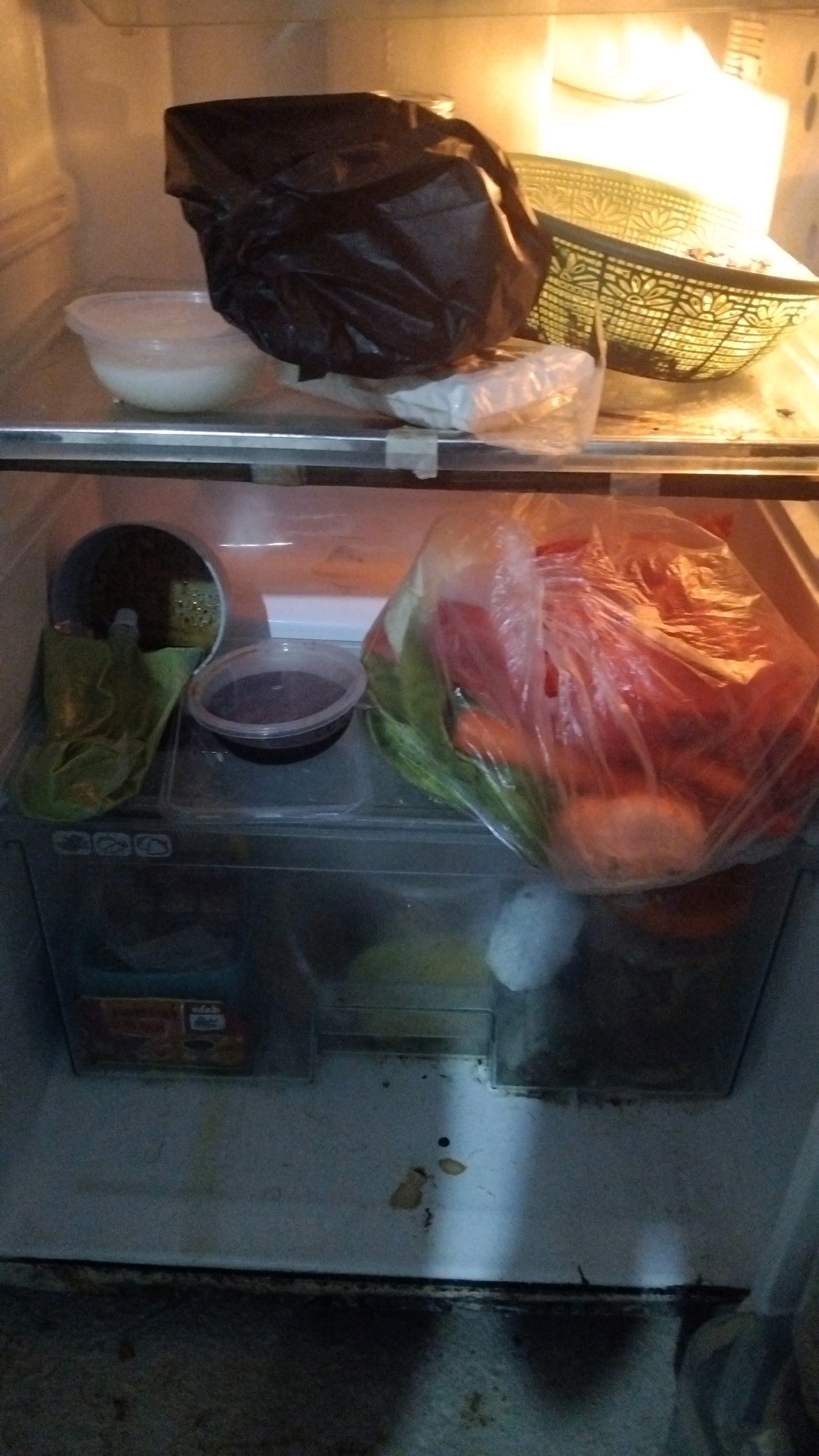 The image shows the interior of a refrigerator in Kebon IX, Indonesia. On the top shelf, a black plastic bag is placed next to a green woven basket. Below, on the second shelf, a clear plastic container with a white substance, likely rice, sits to the left of a large clear plastic bag filled with red vegetables, possibly tomatoes and carrots, and green leafy vegetables. To the right of the white substance container, a small clear plastic bowl contains a dark liquid, possibly soy sauce or a similar condiment. The bottom shelf of the refrigerator appears to be a drawer or compartment. The interior of the refrigerator shows some signs of wear and tear, with some spills and debris visible on the surfaces. The lighting suggests the refrigerator door is open, illuminating the contents.