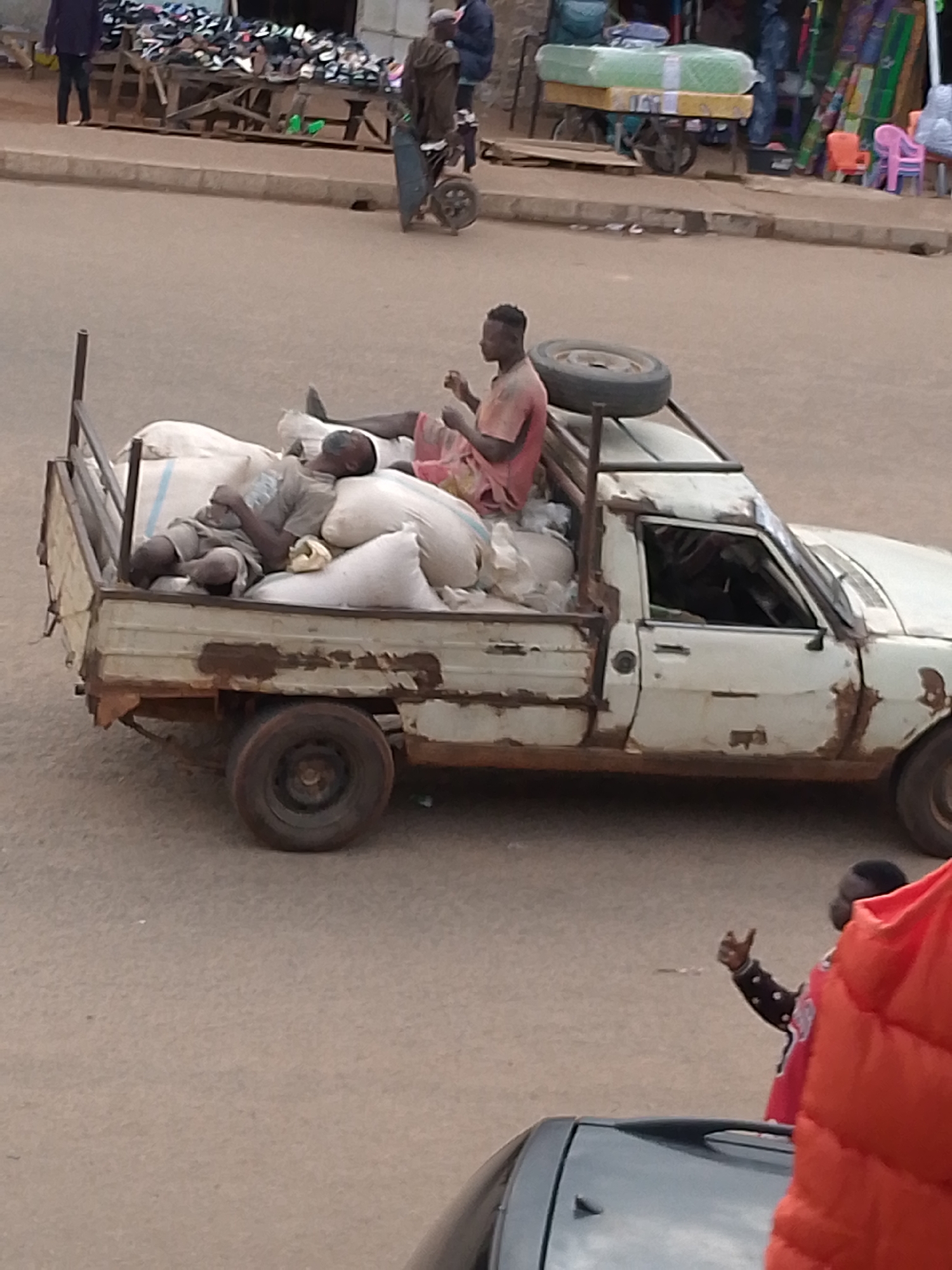 This image depicts a stationary, heavily rusted white pickup truck in a street environment within Bukuru, Nigeria. The truck's cargo bed is filled with numerous white sacks, presumably containing goods. Two adult males are present in the truck bed. One male, wearing a light-colored t-shirt and shorts, is lying down on his side amidst the sacks, appearing to be asleep or resting. The second male, wearing a reddish shirt, is seated upright closer to the cab, holding a small object in his hands. A dark spare tire is secured near the cab in the truck bed. The vehicle exhibits significant rust and wear along its lower body panels and around the doors.
In the background, a raised pavement or curb runs parallel to the road. On the left, a person in dark clothing is walking away from the camera, near a wooden stall displaying numerous shoes. Further into the background on the right, another mobile cart or stall holds a mattress wrapped in plastic, with various colorful items and small plastic chairs (orange and pink) visible behind it. The road surface is light-colored asphalt or dirt. In the immediate foreground, to the bottom right, a person's arm is visible, making a "thumbs up" gesture, with a red garment partially obscuring their body. The overall scene suggests a busy, utilitarian, and possibly market-oriented area.