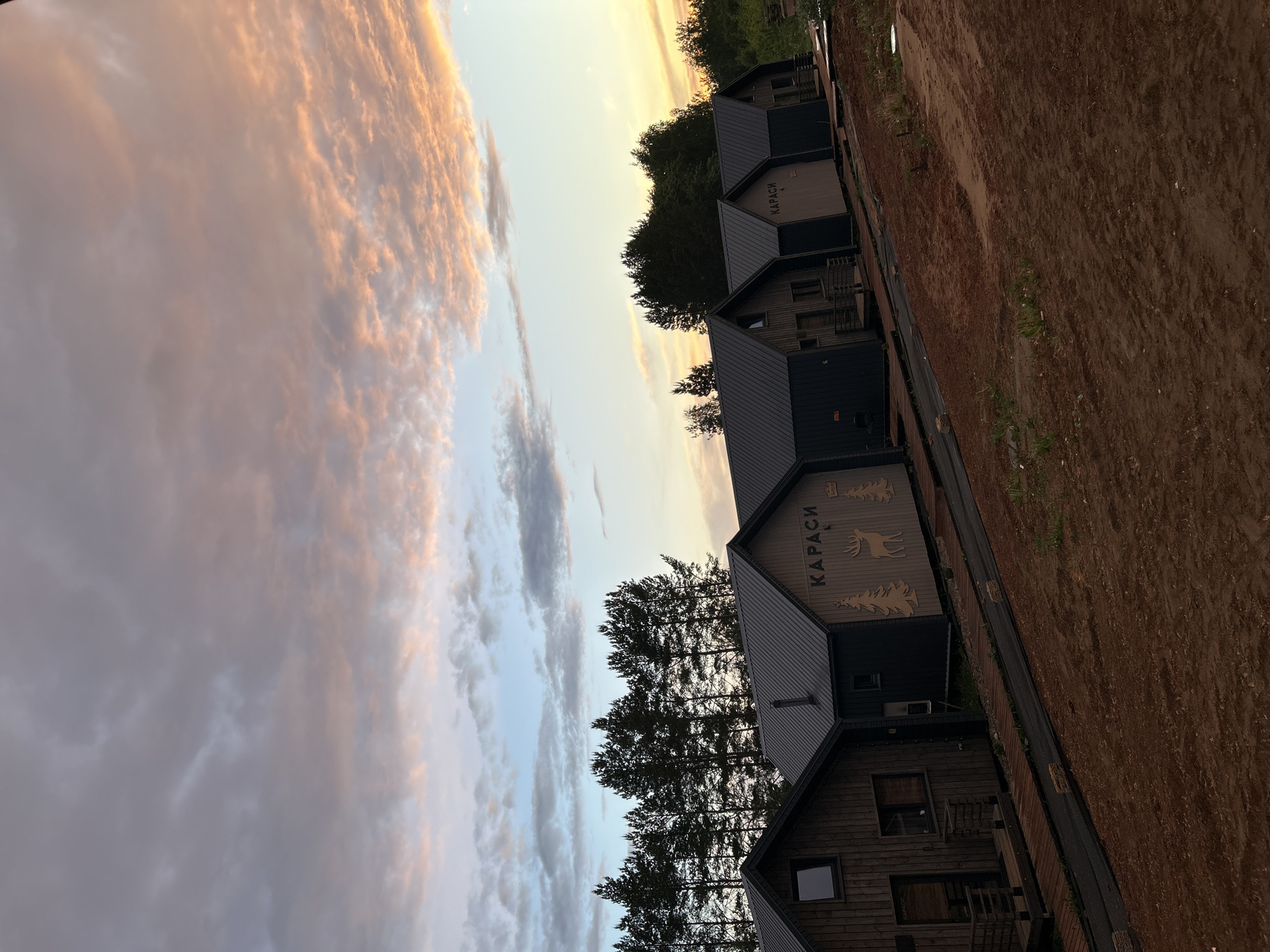 The image shows a row of wooden cabins, likely in a rural or semi-rural setting, under a sky with sunset colors. The cabins have dark roofs and light-colored facades with the word "KARASI" inscribed in a vertical orientation, along with deer and tree graphics. Tall pine trees stand between the cabins. To the right of the cabins is an area of brown, bare earth. The angle of the photograph is slightly askew. While the architecture may be similar to structures found in Pirogovo, Russia, without additional evidence, a definitive location identification cannot be made.