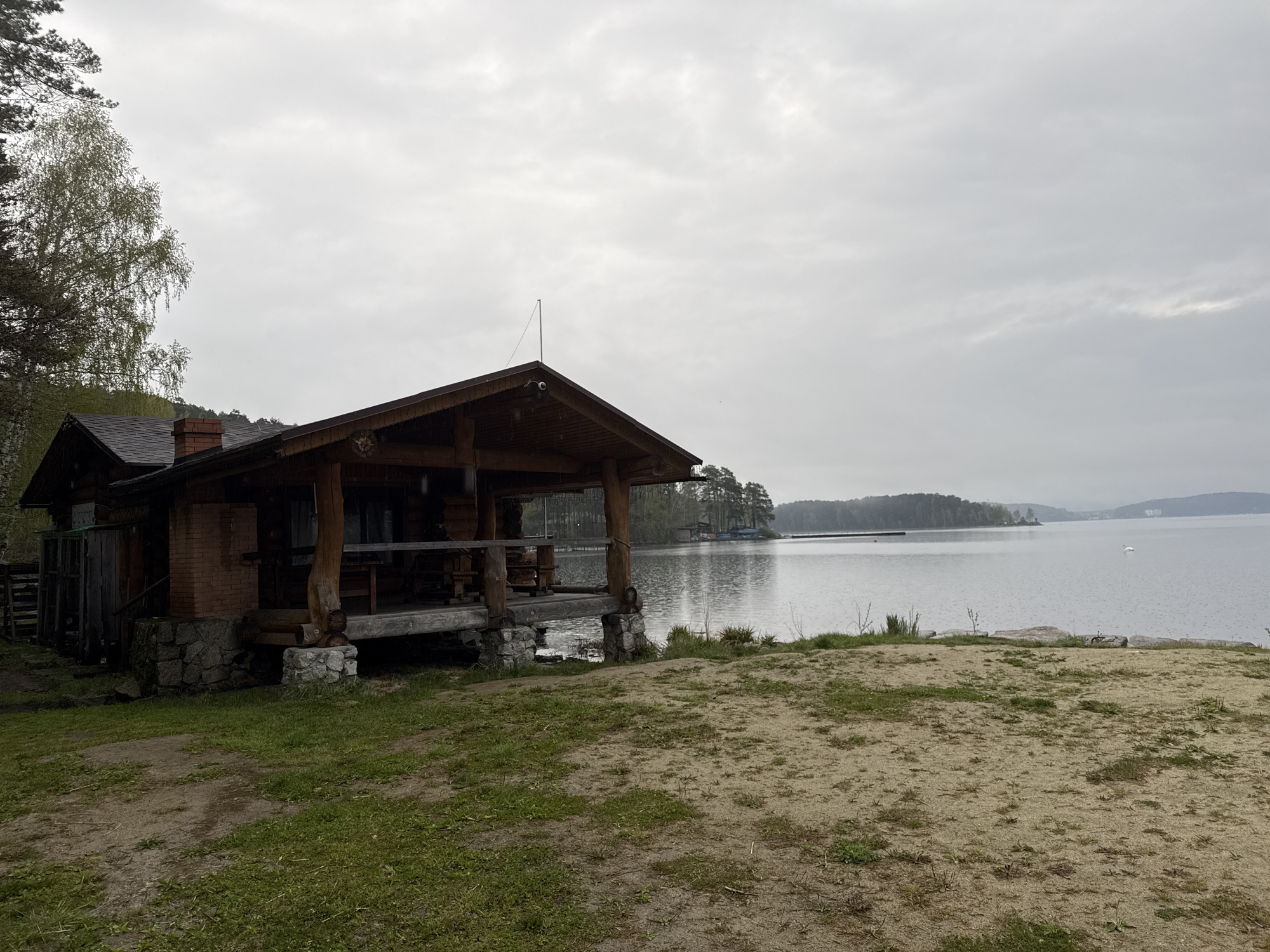 A low-angle, wide shot shows a wooden cabin situated on the shore of a large body of water, possibly a lake or bay. The cabin has a dark, peaked roof with a visible antenna and security camera. It has a porch extending out from the main structure with wooden supports resting on stone bases. The cabin appears to be constructed from logs and brick. The environment surrounding the cabin consists of grassy and sandy areas.
The body of water stretches into the distance, with visible landmasses and a long pier or breakwater. A single white swan floats on the water. The sky is overcast, casting a grey tone over the entire scene. The location is Zolotoi Plyazh, Russia.