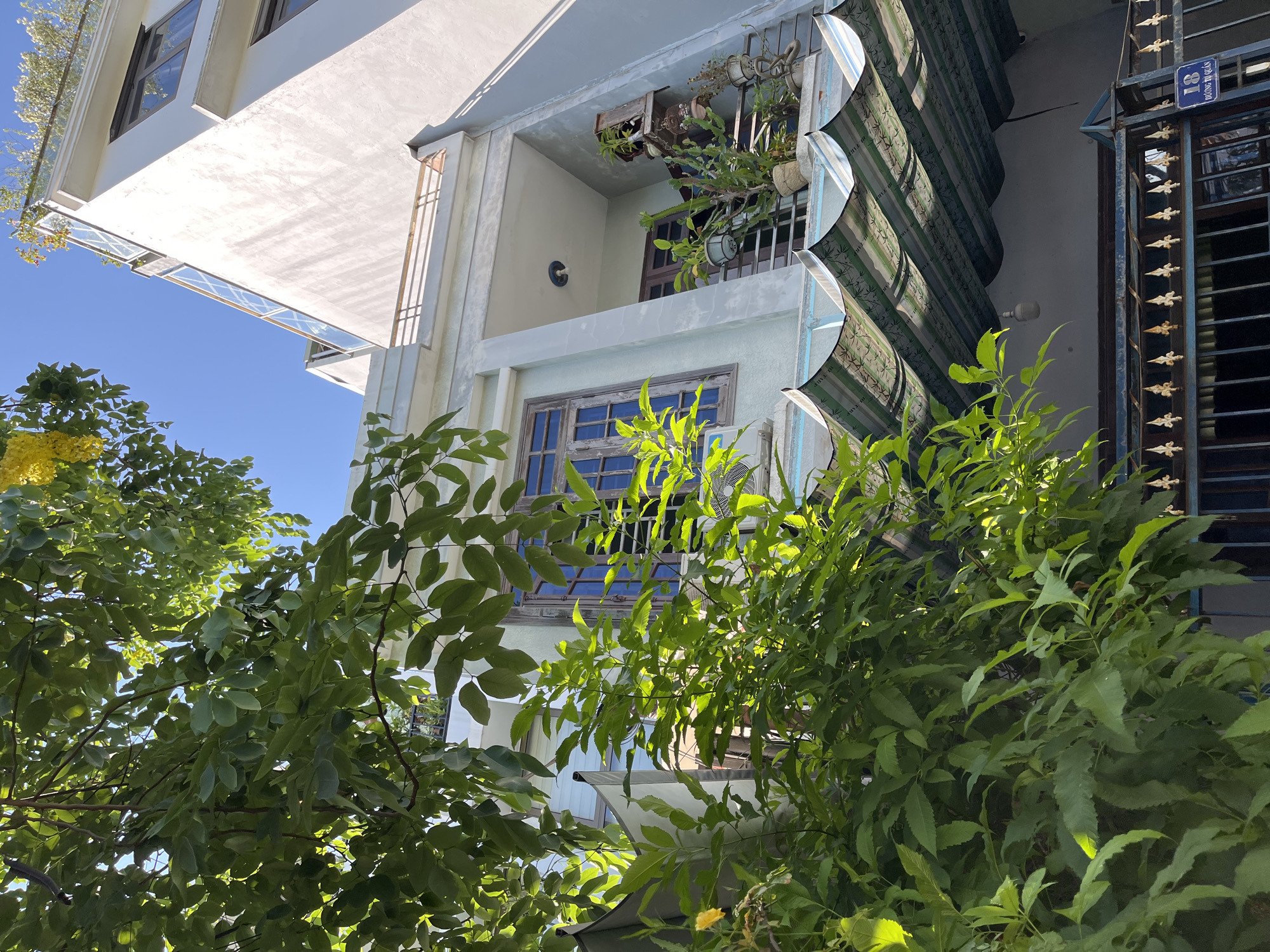 An eye-level, upward-angle shot of a multi-story building framed by green trees and vegetation. The building has white and light blue painted surfaces. A balcony on the second floor is adorned with potted plants. Green and white striped awnings partially cover a section of the building. To the right of the building is a dark blue metal gate with decorative gold accents. A blue street sign reading "18 ĐƯỜNG DỊ QUẦN" is attached to the gate. The sky is visible in the upper left and through gaps in the trees and building. The setting appears to be a residential area in the city of Đà Nẵng, Vietnam, based on the street sign. No people are visible.