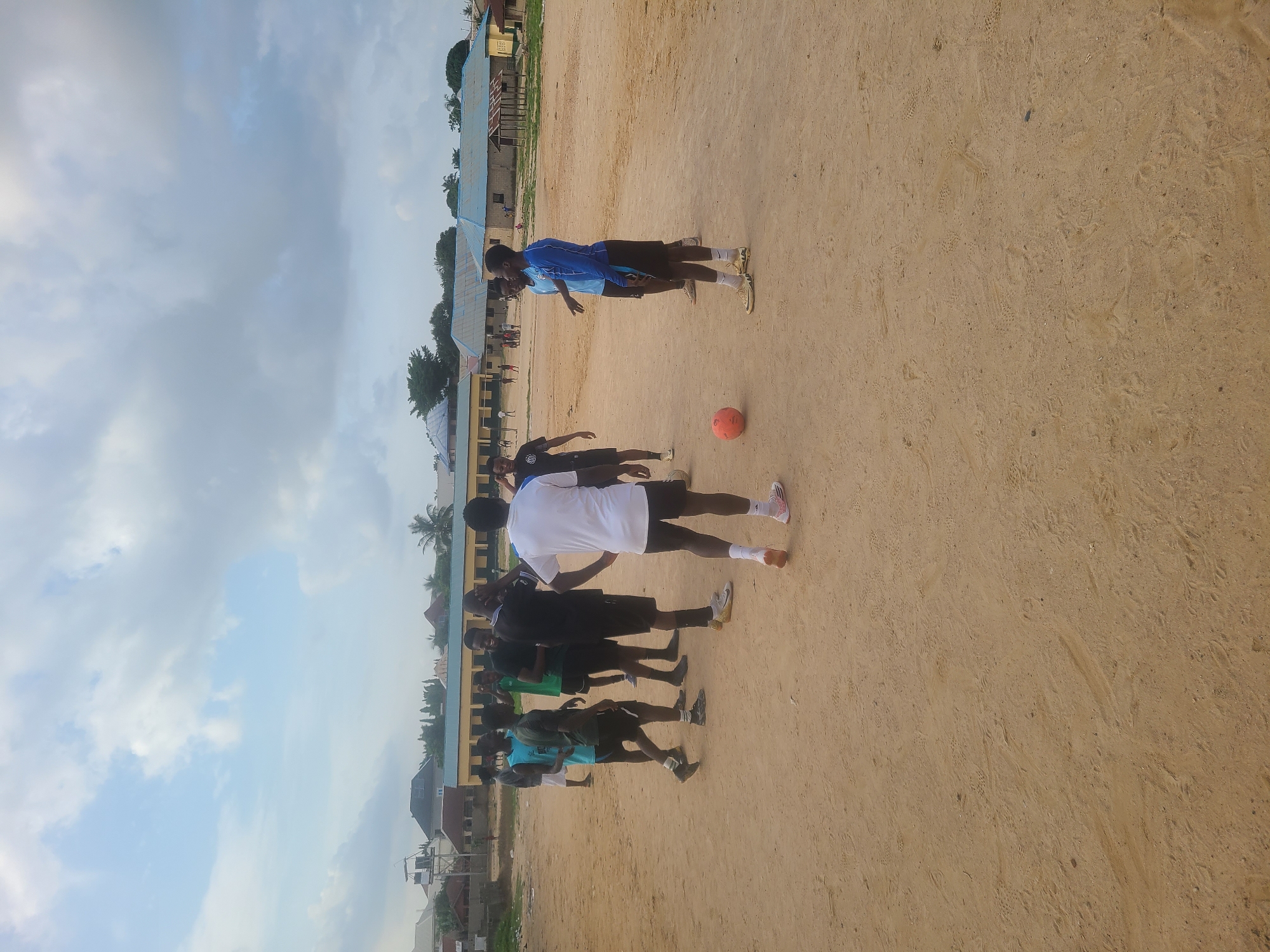 In Masaka, Nigeria, a group of young men are gathered on a dusty, open field under a cloudy sky. They appear to be playing or preparing to play soccer. In the foreground, a bright orange soccer ball rests on the ground. Several individuals are dressed in athletic attire, including t-shirts, shorts, and soccer cleats. Some are standing and observing, while others are engaged in conversation or appear to be mid-action. In the background, modest buildings with corrugated metal roofs are visible, suggesting a semi-urban or rural setting. The overall atmosphere is casual and active, with a sense of community engagement in sports.