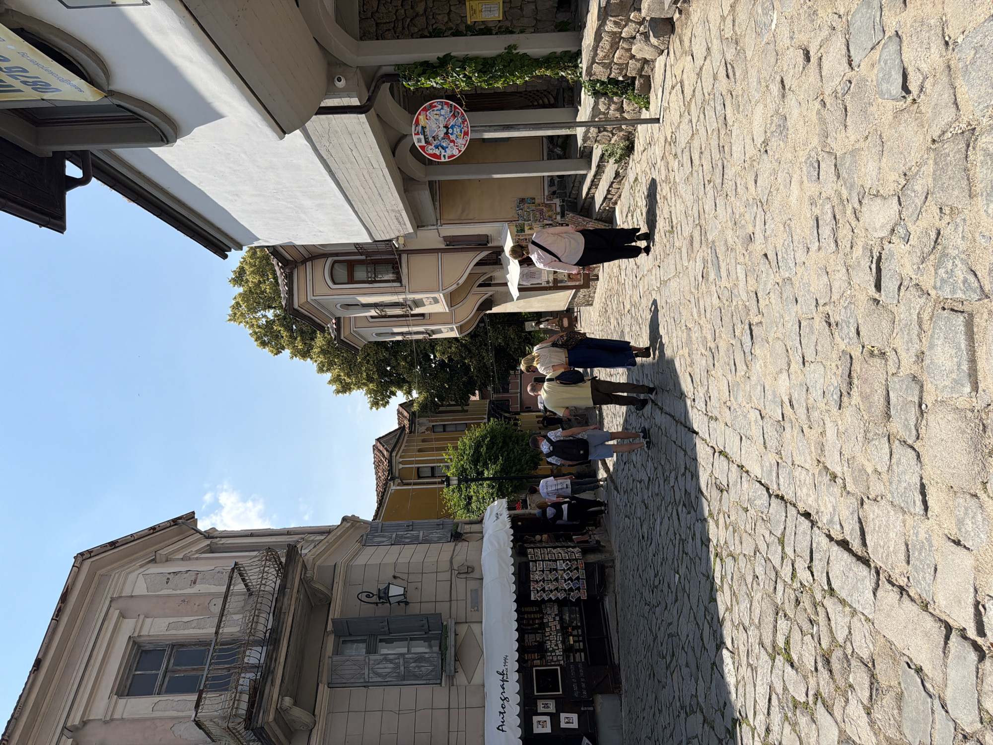 This image captures a bright daylight scene in a narrow, cobbled street within the Old Town of Plovdiv, Bulgaria. The perspective is looking downhill or along a slight curve in the street, with buildings lining both sides. On the left, a two-story building with a light beige, somewhat weathered facade dominates the foreground. Its upper floor features a window with a highly ornate, rusted wrought-iron balcony. Below, a window with closed grey wooden shutters and a black wall-mounted lantern is visible. The ground floor of this building houses a shop with a white scalloped awning, bearing the text "Autograph since 1994." The shop's display window is filled with postcards and various small items. Further down the street on the left, other traditional Plovdiv-style houses with colorful facades and distinct bay windows are visible, interspersed with green trees. On the right side of the street, the foreground building has a white upper section and a rough stone base. A dark brown roof overhang with a gutter system is prominent. A small security camera is mounted on the wall. A banner partially visible at the top edge of this building displays "0876" and "sales@broker.bg," indicating a real estate business. Further down, another building with ornate bay windows, similar to those on the left, can be seen, partially obscured by trees. A green vine grows vertically up the stone wall of the right-side building. A circular red sign, affixed to a metal pole, stands on the right side of the street, covered in various colorful stickers and featuring the word "GAME" at its bottom right. The street itself is paved with uneven, grey and brown cobblestones, with patches of light-colored sand and some small green weeds growing between them. Sunlight casts distinct shadows from the buildings and people onto the street surface. Approximately eight to ten individuals are present in the scene, mostly walking away from the camera's perspective. In the mid-ground, a man in a light yellow t-shirt and dark pants, carrying a black shoulder bag, walks next to a man in a black t-shirt, blue shorts, and a black backpack. Behind them, a woman in a light pink top and black pants, with a dark shoulder bag, walks alongside another woman in a blue skirt and light top, carrying a light bag. Other indistinct figures are visible further down the street. The sky is clear and blue with one small white