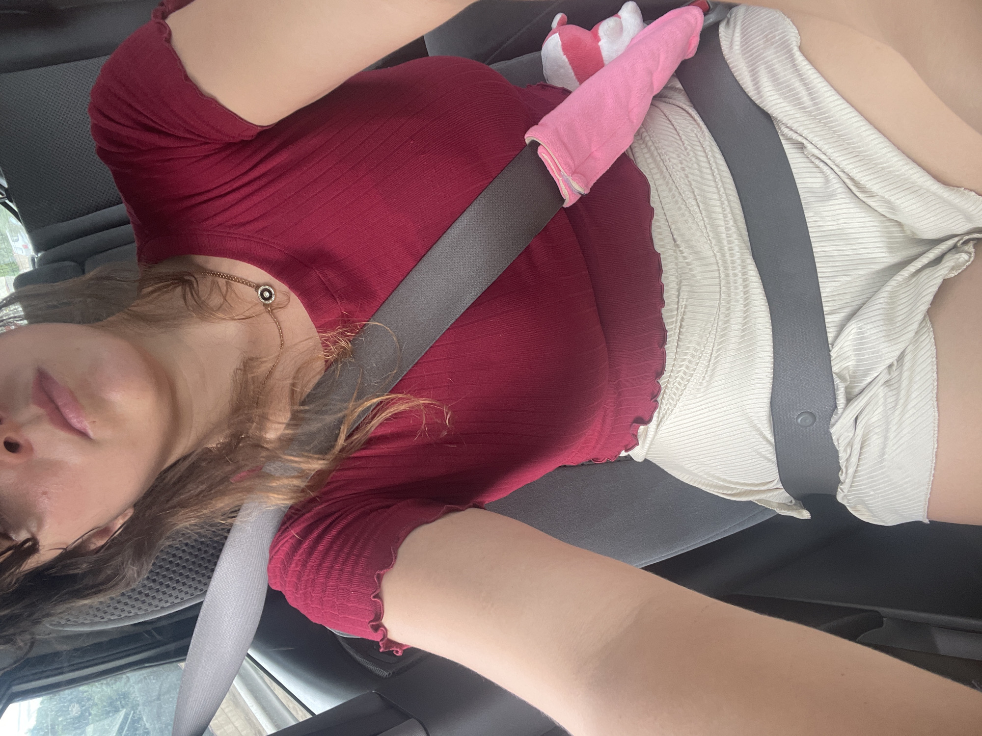 The image shows a close-up, indoor shot of a person sitting in a car, wearing a dark red ribbed top and light-colored ribbed shorts. A gray seatbelt is visible, with a pink padded cover over it. The person has long brown hair and is wearing a necklace. The background consists of car seats and windows, suggesting the car is in motion, possibly on a highway given the blurry exterior visible through the window. The lighting appears to be natural daylight. The specific location in the city of L'Estrella, Spain, cannot be determined from the visual information provided in the image.