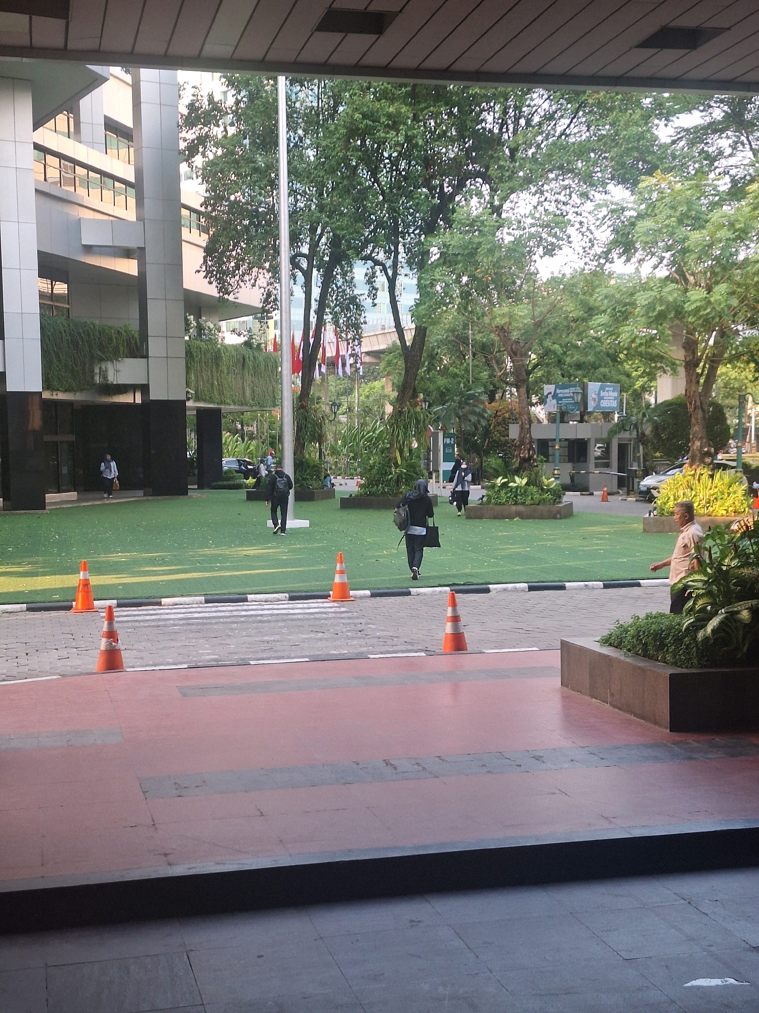The image depicts the exterior of the CIMB Niaga Tower in South Jakarta, Indonesia. The foreground features a red and grey tiled walkway with several orange traffic cones and white crosswalk markings. Beyond this, a meticulously maintained artificial turf area is visible, dotted with trees and foliage. Several individuals are present in the scene, walking across the turf and near the building entrance. One person is entering a building on the left, while others are seen walking on the turf. In the background, the modern architecture of the CIMB Niaga Tower, characterized by its reflective glass windows and geometric design, dominates the scene. The presence of flags and greenery suggests a well-maintained urban environment.