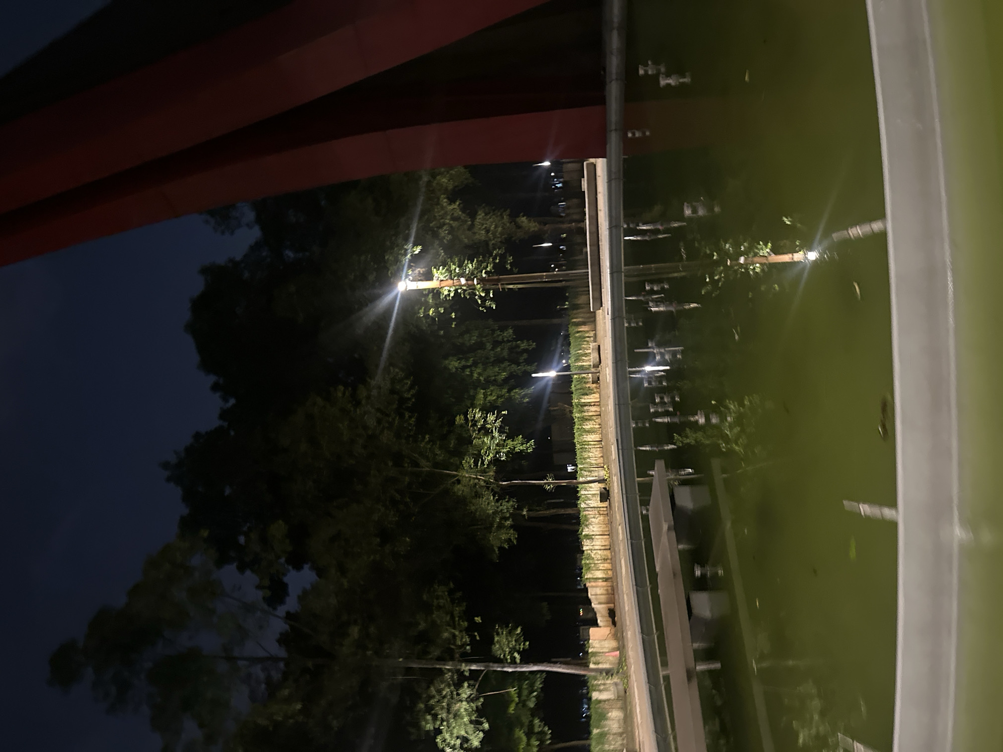 At night in Central Jakarta, Indonesia, the image shows a dimly lit area with trees and lights. The scene is viewed from an elevated position, possibly a building or structure, with red and white structural elements visible in the frame. The area appears to be a park or landscaped space, with trees lining a pathway illuminated by streetlights. A body of water reflects the lights and surrounding environment.