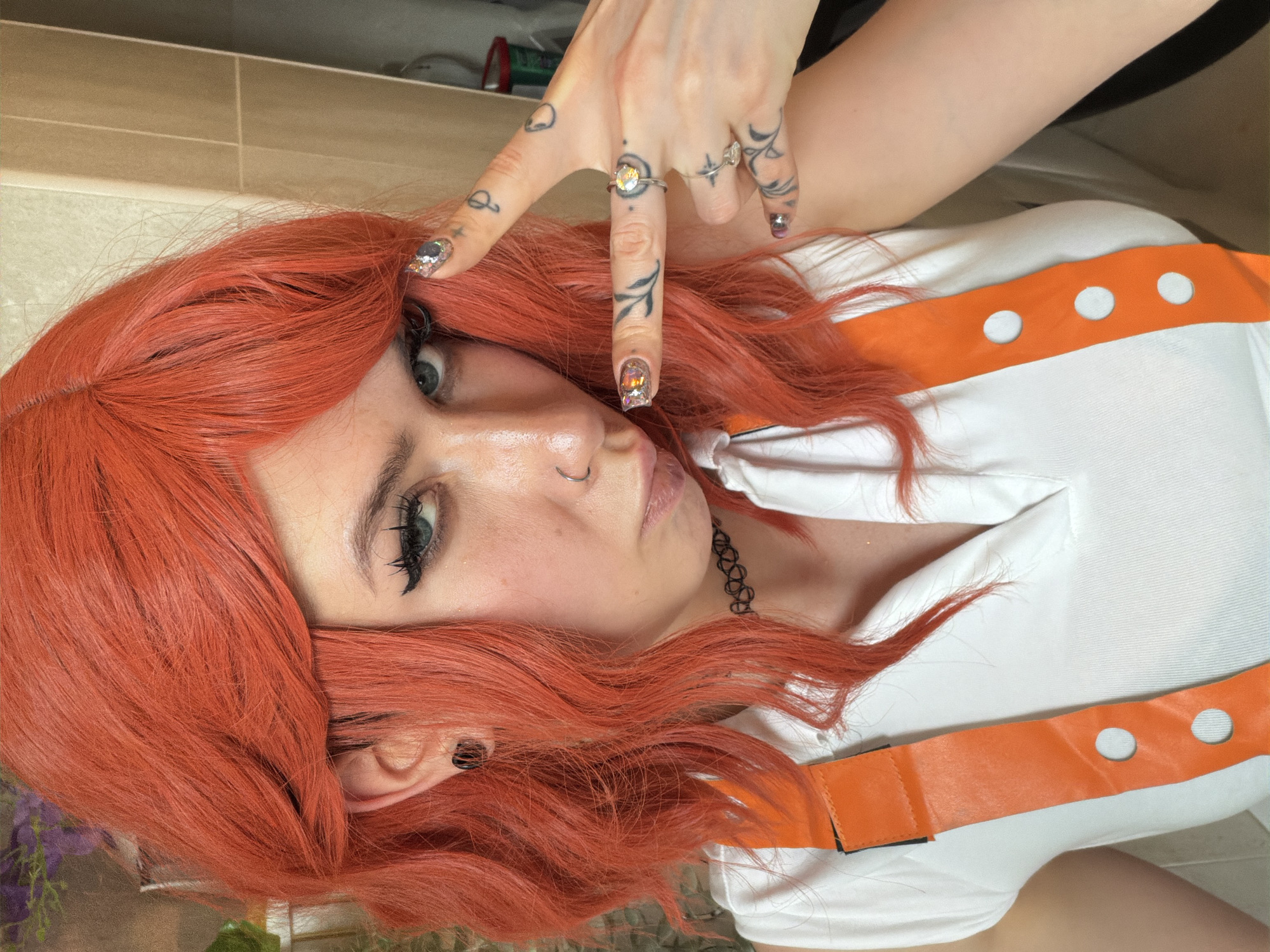 An upward-facing, vertically oriented close-up shot features a person with vibrant, wavy, orange-red hair, likely a wig, lying down indoors. The subject has fair skin, light-colored eyes (blue-green), and is looking directly at the camera with a slightly pouting expression. Heavy black eyeliner and mascara accentuate the eyes, and a small silver hoop nose ring is visible. A black choker with an intricate cutout design is around the neck.
The person is wearing a white top or shirt, partially covered by an orange strap or harness with three white circular cutouts, suggesting a costume or uniform. One arm is raised, with the hand positioned near the head, fingers splayed into a "V" sign. This hand displays multiple small, intricate tattoos on the fingers and the back of the hand, including designs like an alien head, a crescent moon, and leafy patterns. Several rings adorn the fingers, notably one with a large yellowish gemstone on the index finger. The fingernails are elaborately decorated with glittery, jewel-toned polish.
The background shows light beige or tan tiled walls, indicating an indoor setting, possibly a bathroom or a similar tiled space. Hints of what appear to be purple foliage or flowers are faintly visible in a reflection or in the deeper background in some crops. The image captures a casual, self-aware pose within an interior space in Budva, Montenegro. There is no visible text in the image.