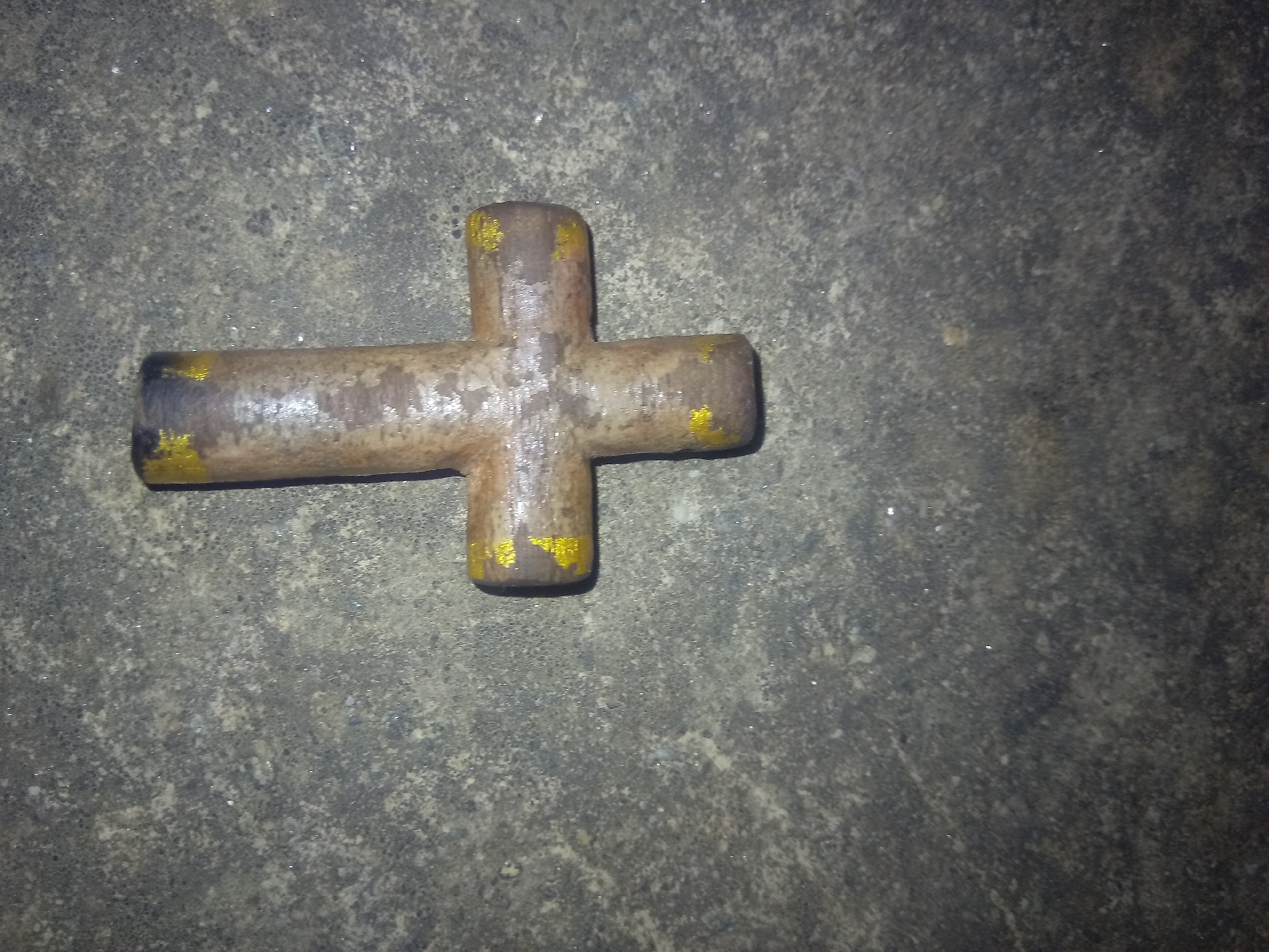 A rusty, possibly metallic, cross-shaped object lies on a textured, gray surface that appears to be concrete or asphalt. The cross has a cylindrical shape and shows signs of wear and oxidation, with patches of a faded, possibly yellow, paint visible on its extremities. The gray surface is uneven and speckled with darker and lighter tones, with some areas appearing smoother than others. There are no other objects or discernible features in the frame. No location is indicated.