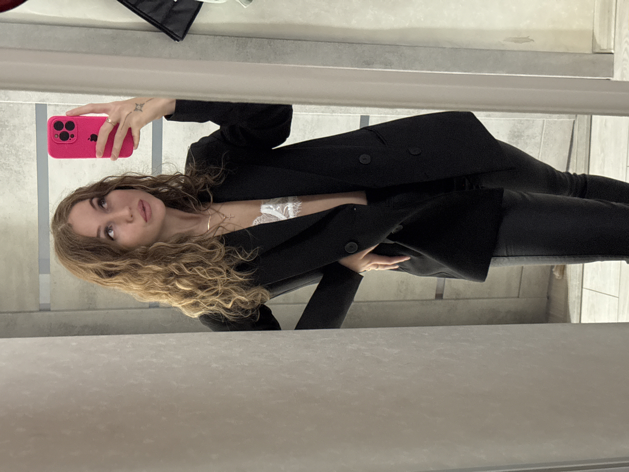 The image is a mirror selfie taken by a woman in Chaikovsky, Russia. The primary subject is a young woman with long, voluminous, curly light brown hair that flows over her shoulders. She is looking upwards and to the side, directly at her reflection in the mirror, with a contemplative or poised expression.
She is dressed in a chic, semi-formal outfit featuring an open black double-breasted blazer with two prominent buttons visible on the right lapel area. Beneath the blazer, she wears a delicate white lace top or bralette, with intricate patterns visible on the fabric. Her lower attire consists of black pants, completing a monochromatic ensemble.
In her left hand, which is held up to capture the photo, she is holding a bright pink smartphone. The phone case has a textured surface, and three distinct camera lenses are visible on its back. A small, star-shaped tattoo is visible on the back of her left hand, just above her wrist, and she wears a subtle ring on her left ring finger. A thin gold chain necklace with a small cross pendant adorns her neck.
The setting is an indoor space, reflected in the mirror. The background shows light-toned, possibly tiled or paneled walls, with a distinct horizontal line running through the mid-ground, which could be a shelf, counter, or architectural detail. The lighting appears to be ambient indoor light. There is no visible text in the image.