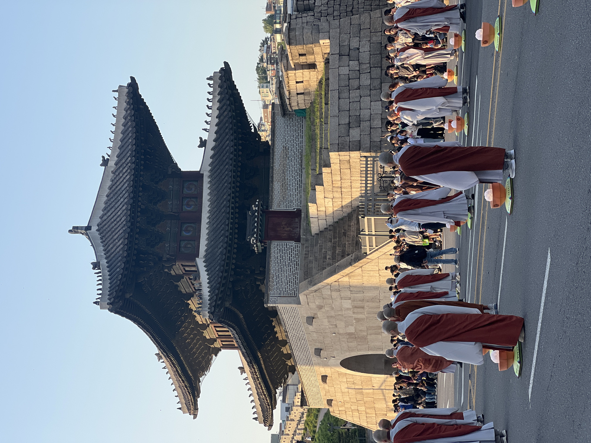 The image shows a solemn procession in front of Namdaemun Gate, a prominent historic gateway in Seoul, South Korea. A line of individuals wearing monastic robes are bowed down. Behind them, a larger crowd of onlookers is visible. The foreground is dominated by the modern asphalt road marked with white and yellow lines, contrasting with the historic stone structure of the gate and surrounding wall. The sky is light blue, suggesting either early morning or late afternoon lighting conditions. The scene captures a blend of tradition and modernity, with the historic gate serving as a backdrop for a ritualistic event.