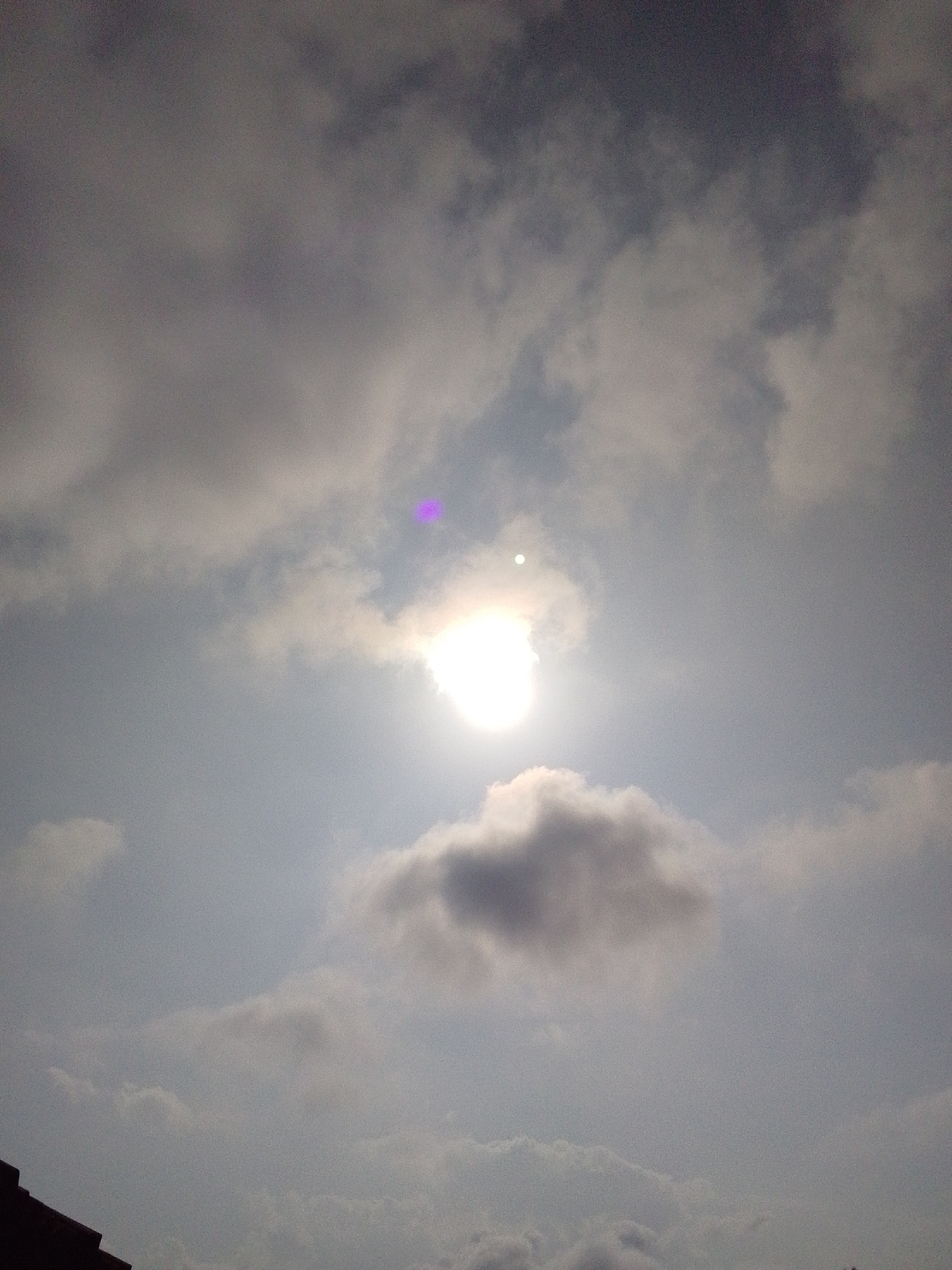 The image depicts a cloudy sky with the sun partially obscured by clouds, creating a bright halo effect. There is a distinct purple lens flare in the upper left quadrant of the image. The lower left corner shows a portion of a dark, angled structure, possibly a roofline or building edge, which is out of focus. The overall scene is dominated by varying shades of gray and white clouds, with the sun being the brightest element. The setting appears to be outdoors during the daytime.