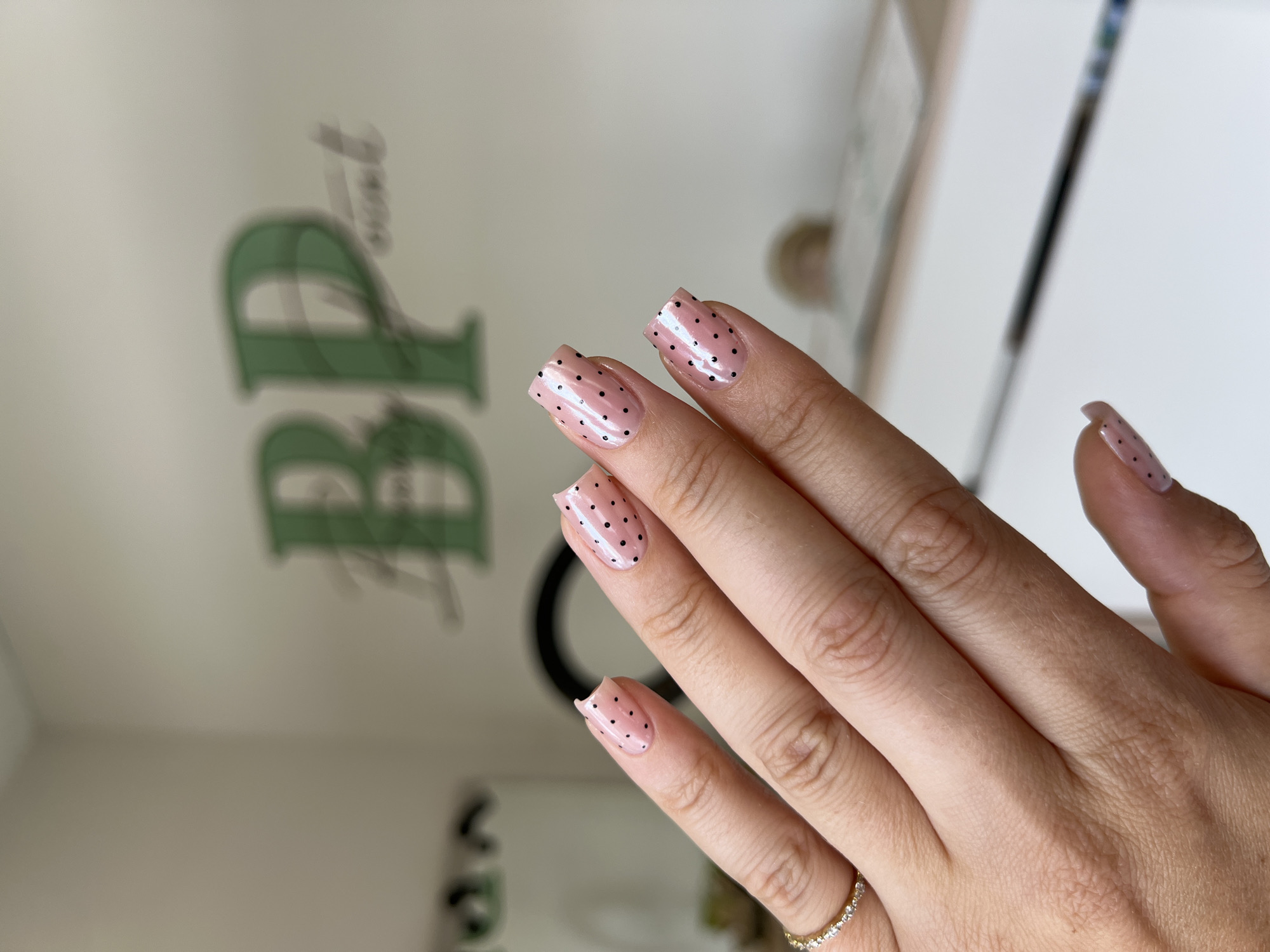 The image shows a woman's hand with light pink nails adorned with small black polka dots. The hand is facing towards the left of the frame, with the fingers slightly curved. A gold ring with small stones is visible on the ring finger. The background includes a blurred image of green letters and a white surface, and it is not possible to identify this location with certainty as Podgorica, Montenegro.
