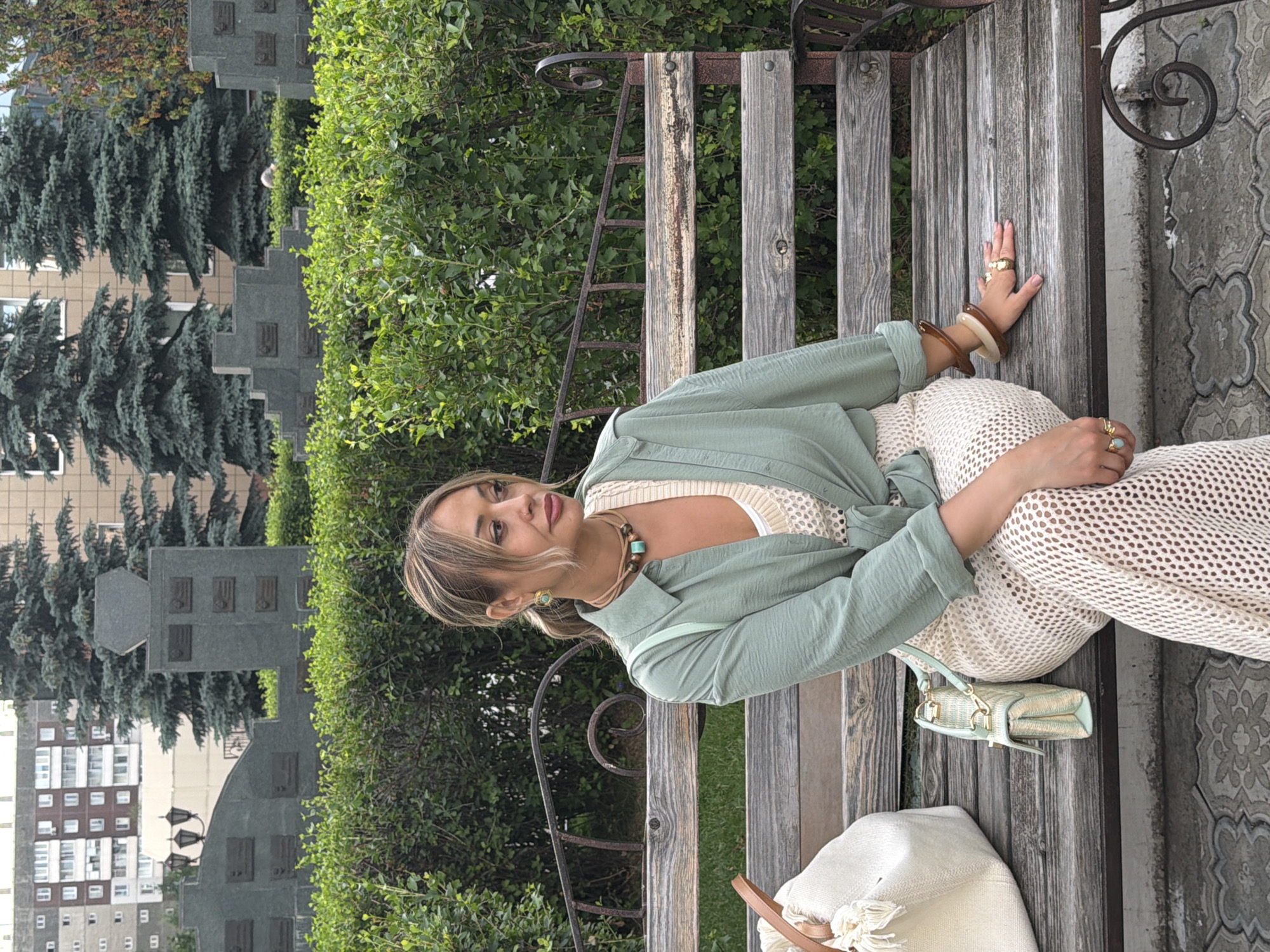 A woman with blonde hair is sitting on a wooden bench outdoors. She is wearing a light green shirt over a cream-colored top and a cream-colored skirt. She has several rings on her fingers and bracelets on her wrists, along with a necklace. A small light green purse is next to her on the bench, and a white bag with tassels is on the ground near her feet.
The background includes lush green bushes and trees, with buildings and a monument visible behind them. The monument appears to be a grey structure with several square reliefs on it, and behind that, there are apartment buildings with windows.
The lighting suggests it is daytime. The woman's expression is relaxed and she is looking towards the camera. The scene appears to be a park or public space.