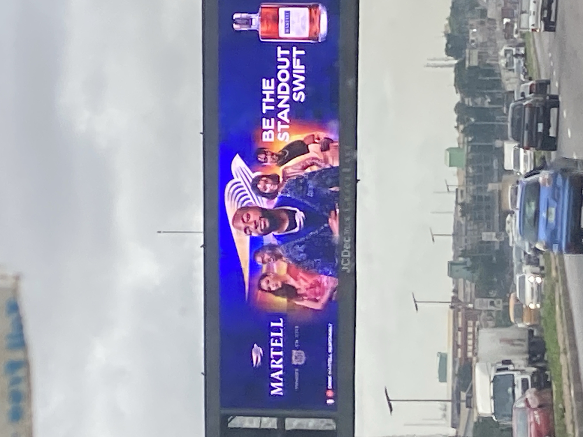 This is a photograph of a billboard advertisement for Martell cognac, situated above a highway with moderate traffic in Ikeja, Nigeria. The billboard features a group of individuals and a bottle of Martell cognac. The text on the billboard reads "BE THE STANDOUT SWIFT". The advertisement is positioned against a cloudy sky. The highway below is populated with various vehicles, including cars, trucks, and vans. The surrounding environment appears to be urban, with buildings and infrastructure visible in the background. The overall impression is a typical scene of urban life and commerce in Ikeja.
