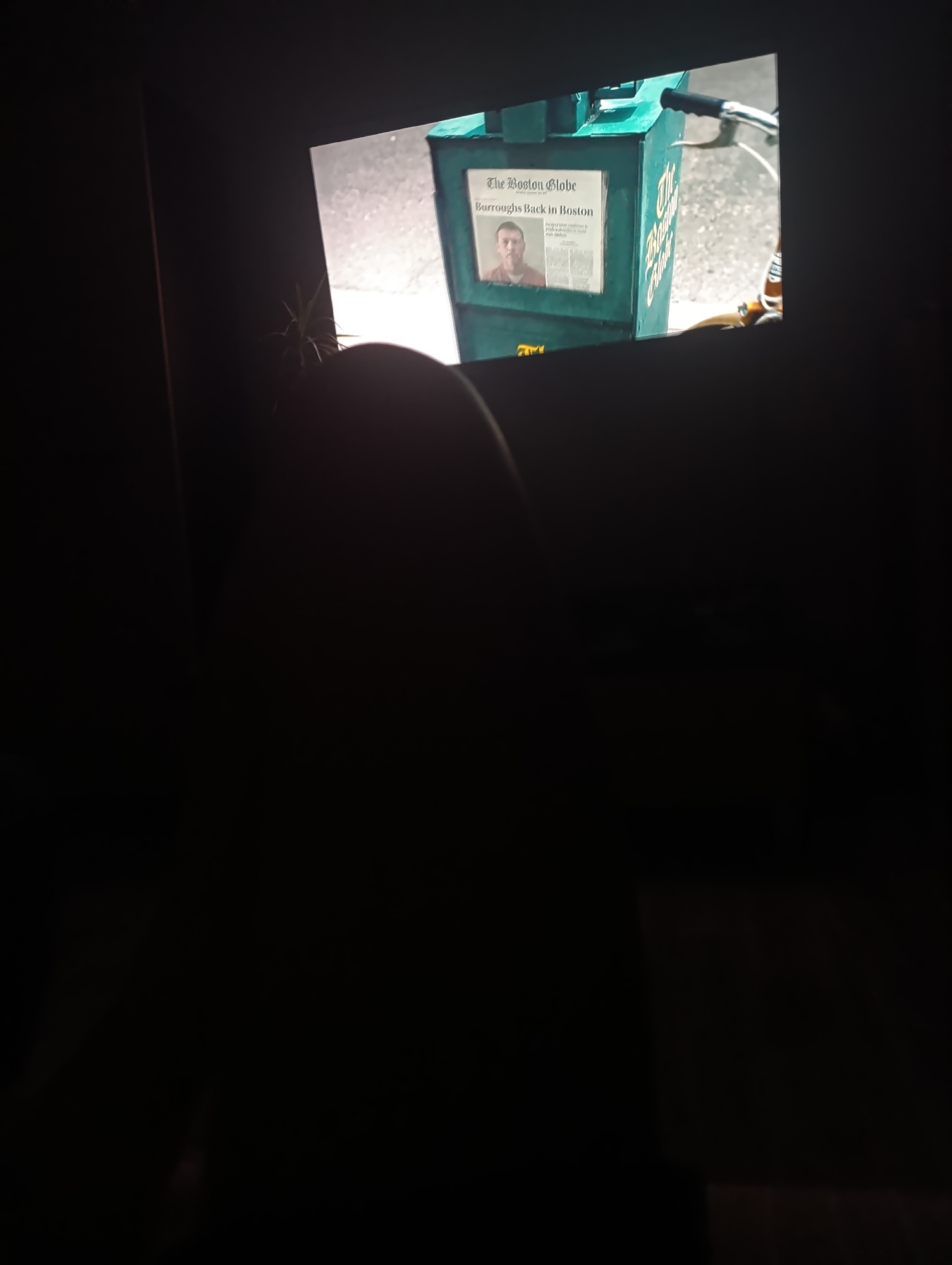 In an indoor setting, a close-up shot displays a television screen displaying a newspaper headline that reads "The Boston Globe Burroughs Back in Boston." The newspaper is situated in a green newspaper stand. A portion of a person's head, appearing to be a shoulder and head, is visible in the foreground, partially obscuring the lower part of the screen. The environment is dark, with the television screen being the primary light source. The background outside the screen is indistinguishable due to the low light conditions. The image's composition suggests a viewing of a screen, possibly a movie or television show.