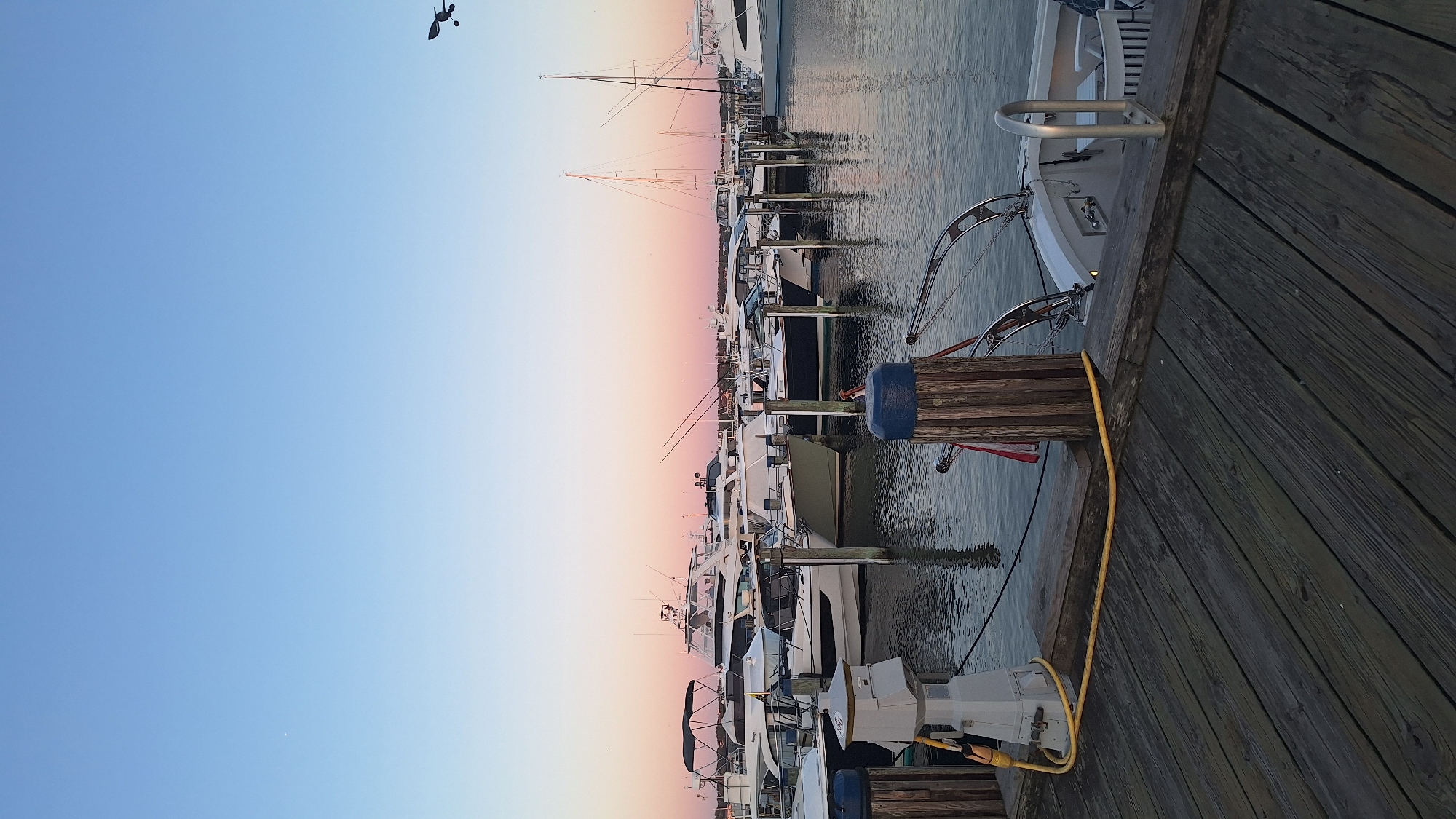 The image depicts a serene marina scene at dusk in Nantucket, United States of America. The sky displays a gradient of soft blues and pinks, indicative of sunset. Several docked boats, ranging from yachts to smaller vessels, are visible. The foreground features a wooden pier with weathered planks, a yellow electrical cord, and wooden posts with blue caps. Reflections of the boats and the sky are visible on the calm water. In the distance, other boats and structures of the marina are present. The overall atmosphere is peaceful and tranquil.