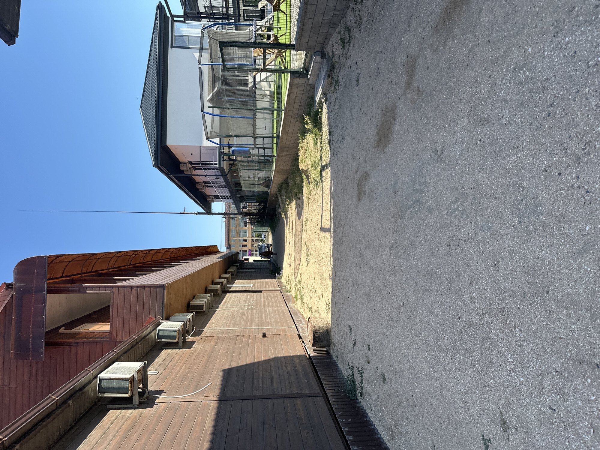 A low-angle, exterior, eye-level shot captures a narrow dirt path between two buildings under a clear blue sky. The building on the left is a multi-story wooden structure with several air conditioning units mounted on its exterior. The building on the right is a white, two-story structure with balconies and an attached trampoline enclosure. A tall, thin antenna stands next to the white building. In the distance, a dark-colored car is visible on the path, with a person walking behind it. Further down the path, a modern multi-story building with balconies and windows can be seen. The ground is covered in dirt and small stones, with patches of dry grass. The scene appears to be in a residential or resort area, possibly in Golubitskaya, Russia, given the architectural style and vegetation.