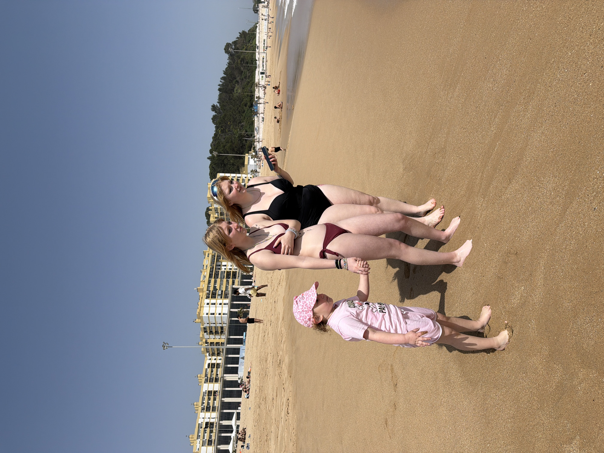Two women are lying on the sandy beach of Praia de Carcavelos, Oeiras, Portugal. One woman wears a black swimsuit and holds a smartphone. The other wears a maroon bikini. A young child in a pink outfit and patterned hat stands holding the second woman’s hand. In the background are buildings and people on the beach near the water’s edge. The sky is blue.