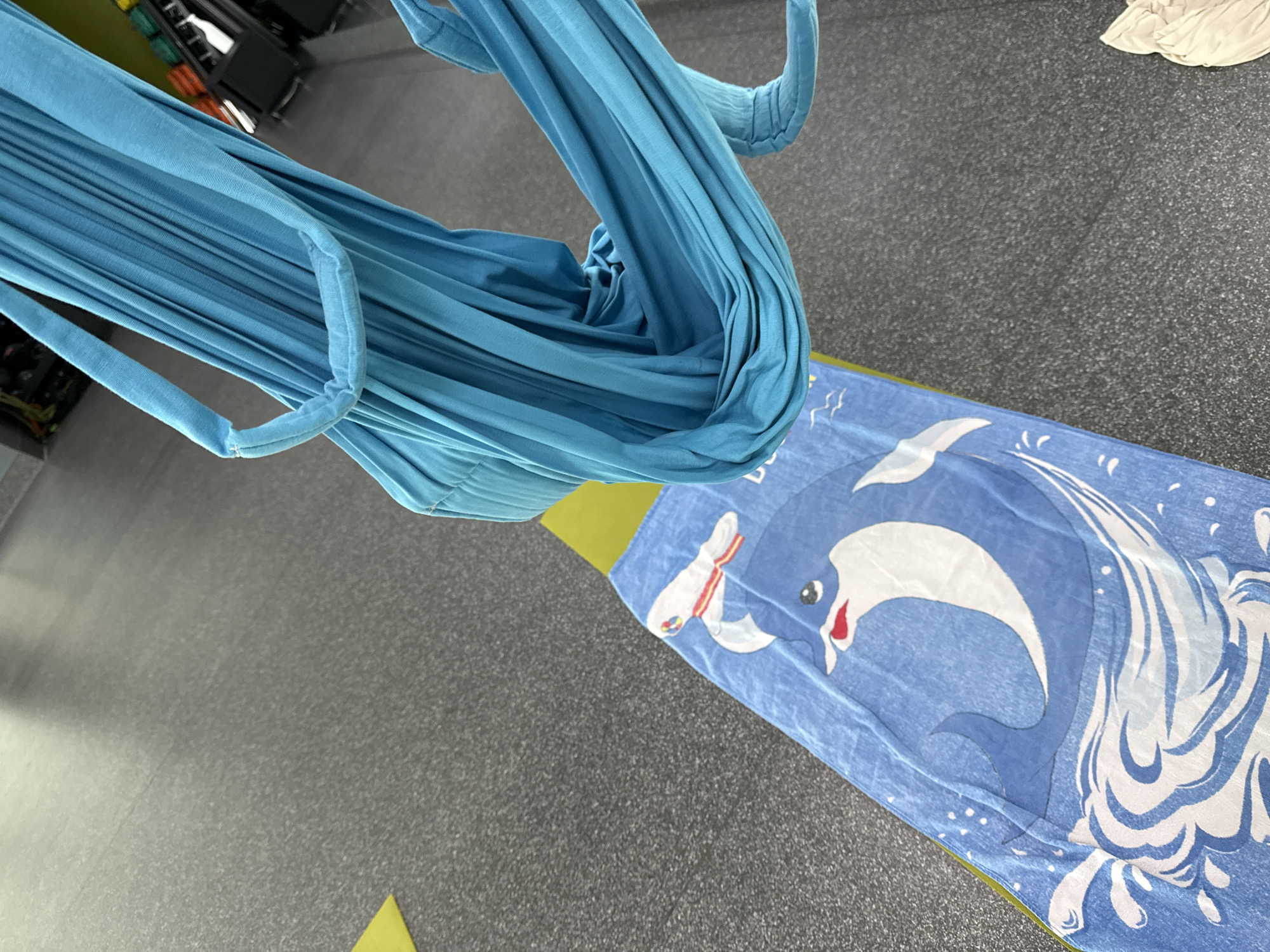 In a gym, a blue aerial yoga hammock hangs from above. Below the hammock, a blue towel with a dolphin design is laid out on a green yoga mat. The floor is a dark gray speckled surface. In the background, weight racks and gym equipment are visible.