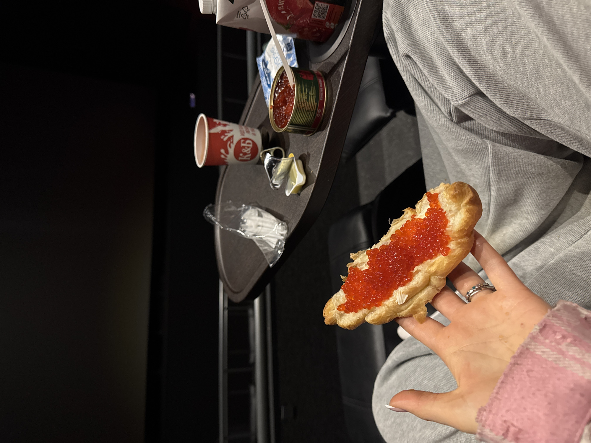 Inside a dark movie theater in Moscow, Russia, a person holds a piece of bread topped with red caviar. A metal tray holds a partially opened can of caviar, a disposable cup from the “K&B” store, small plastic packets, and a white beverage container. The person is wearing a light gray sweatshirt with a glimpse of a pink and white plaid sleeve, and has a ring on their ring finger. The background is dimly lit, showing the texture of the movie theater seats.
