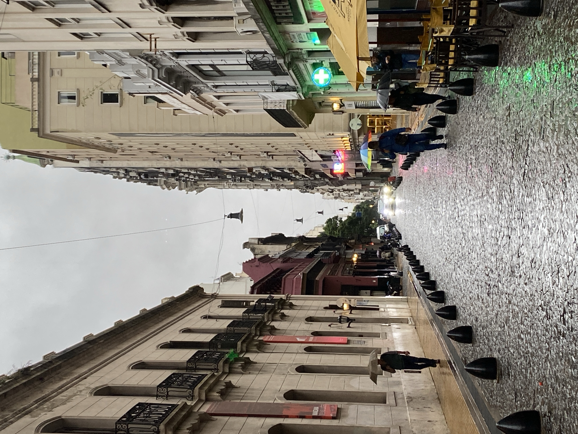 This is a street scene in Buenos Aires, Argentina, likely in the historic district. The image is taken from a pedestrian perspective, showing a narrow street lined with buildings of varying heights and architectural styles. The street is paved with a cobblestone-like material, and it appears to be wet, reflecting light from the surrounding buildings and streetlights. Several people are walking along the street, some with umbrellas, indicating that it is raining or has recently rained. There are also cafe or restaurant tables and chairs visible along the side of the street. The buildings show evidence of European-style architecture, with balconies and decorative details. The sky is overcast.