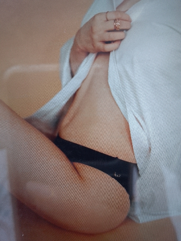The image displays a partial, close-up view of a human figure in an indoor setting. The location is Unknown, Unknown. The figure, identified as female, exhibits light-toned skin. She is partially clad in an unbuttoned or open white collared shirt and black underwear, specifically a brief or thong style garment with a visible label. The subject's left hand, adorned with at least one ring on the ring finger, is positioned near the torso, holding the white shirt open. A portion of a leg is visible in the lower left of the frame. The background is a plain, light beige or peach color, suggesting a wall or fabric backdrop. Diagonal line artifacts are present across the entire image.