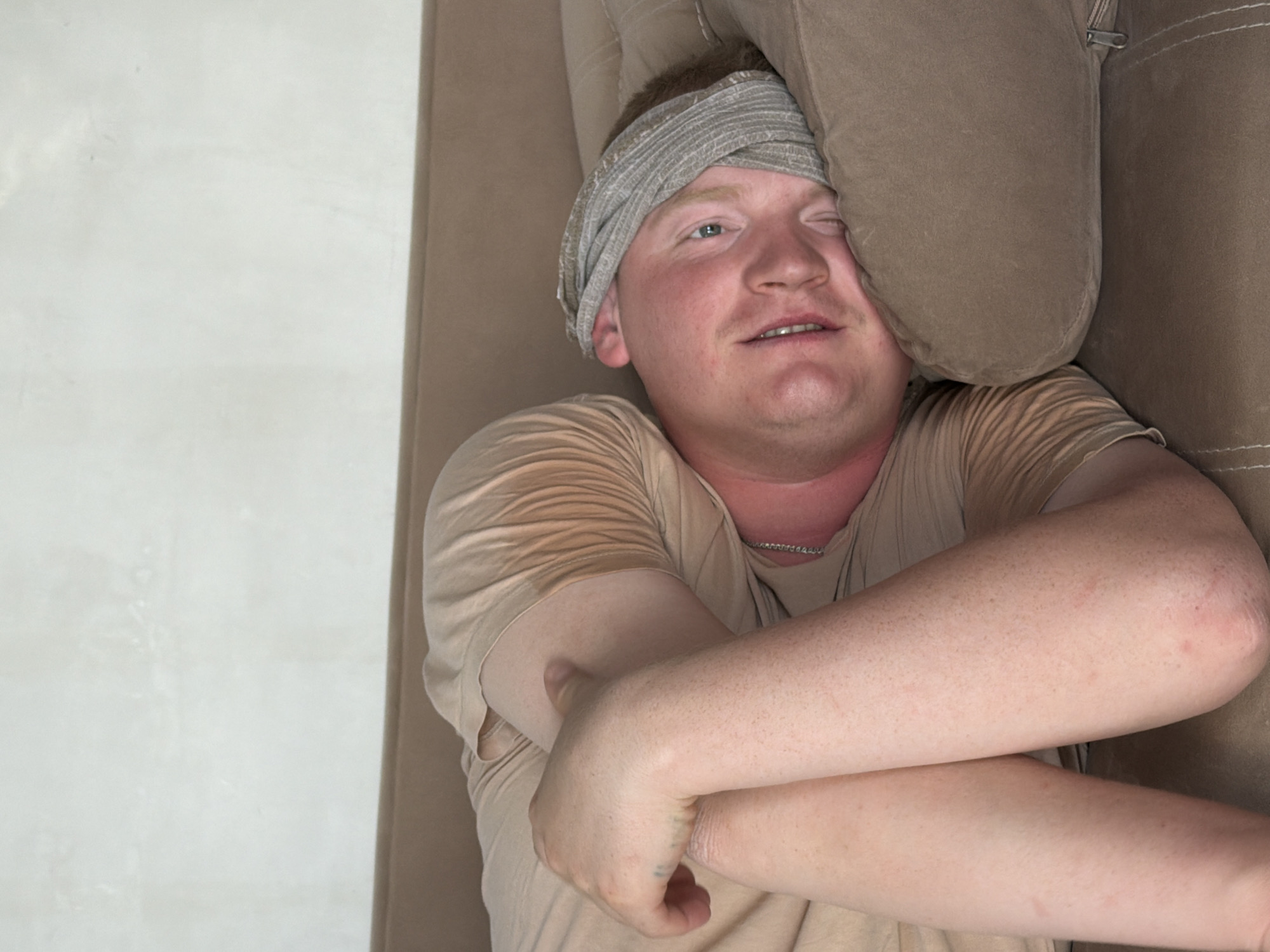 A fair-skinned man is lying on a brown sofa with a gray cloth wrapped around his head. His arms are crossed over his chest. The location is likely indoors. The city of Chaikovsky, Russia, cannot be determined from this image.