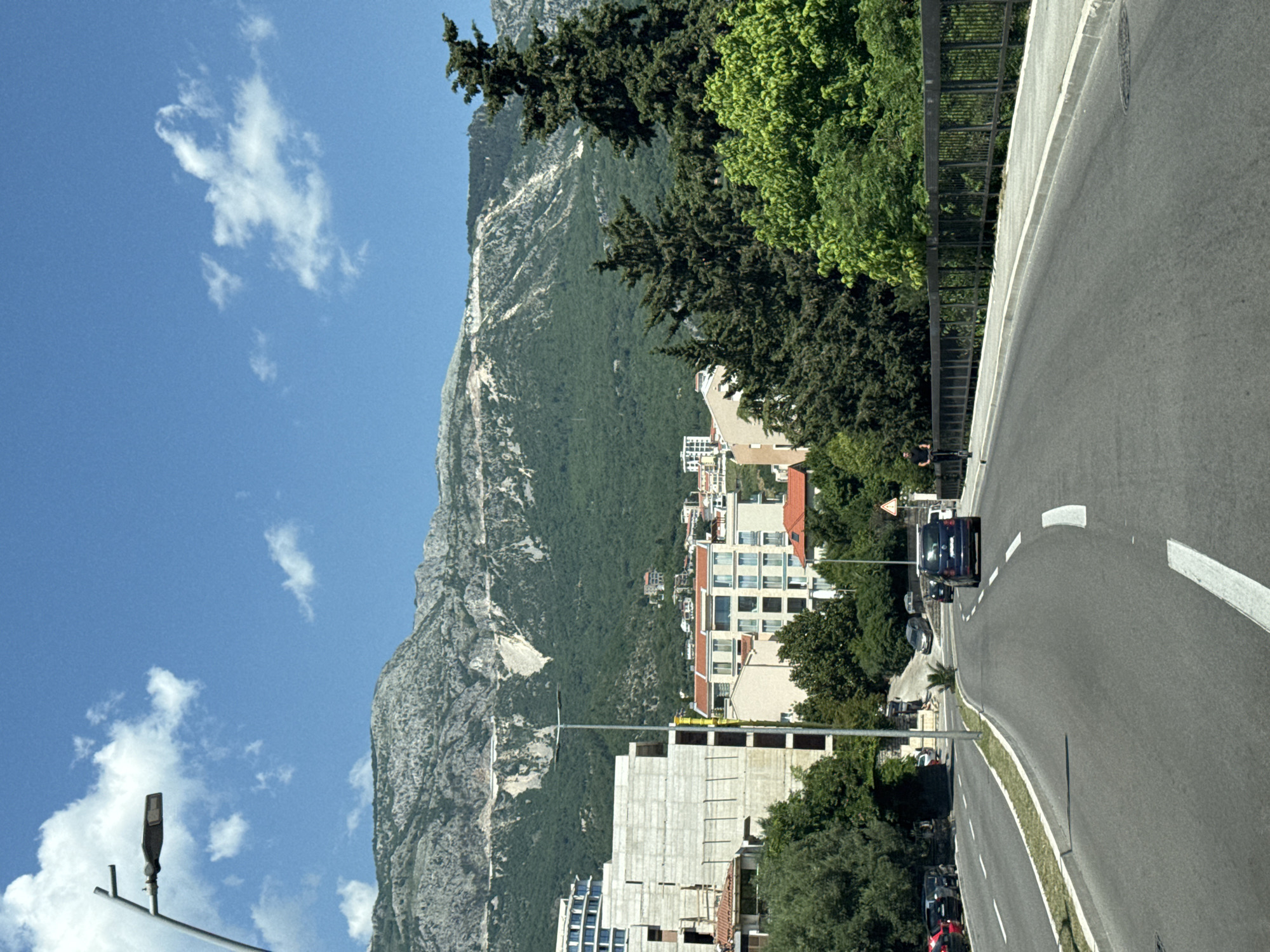 The image depicts a road in Boreti, Montenegro, with a mountain backdrop to the left. Buildings are situated between the road and the mountain. The sky is blue with scattered clouds. Several cars are on the road. A streetlight pole is visible on the left side of the frame. Trees and vegetation line parts of the road.