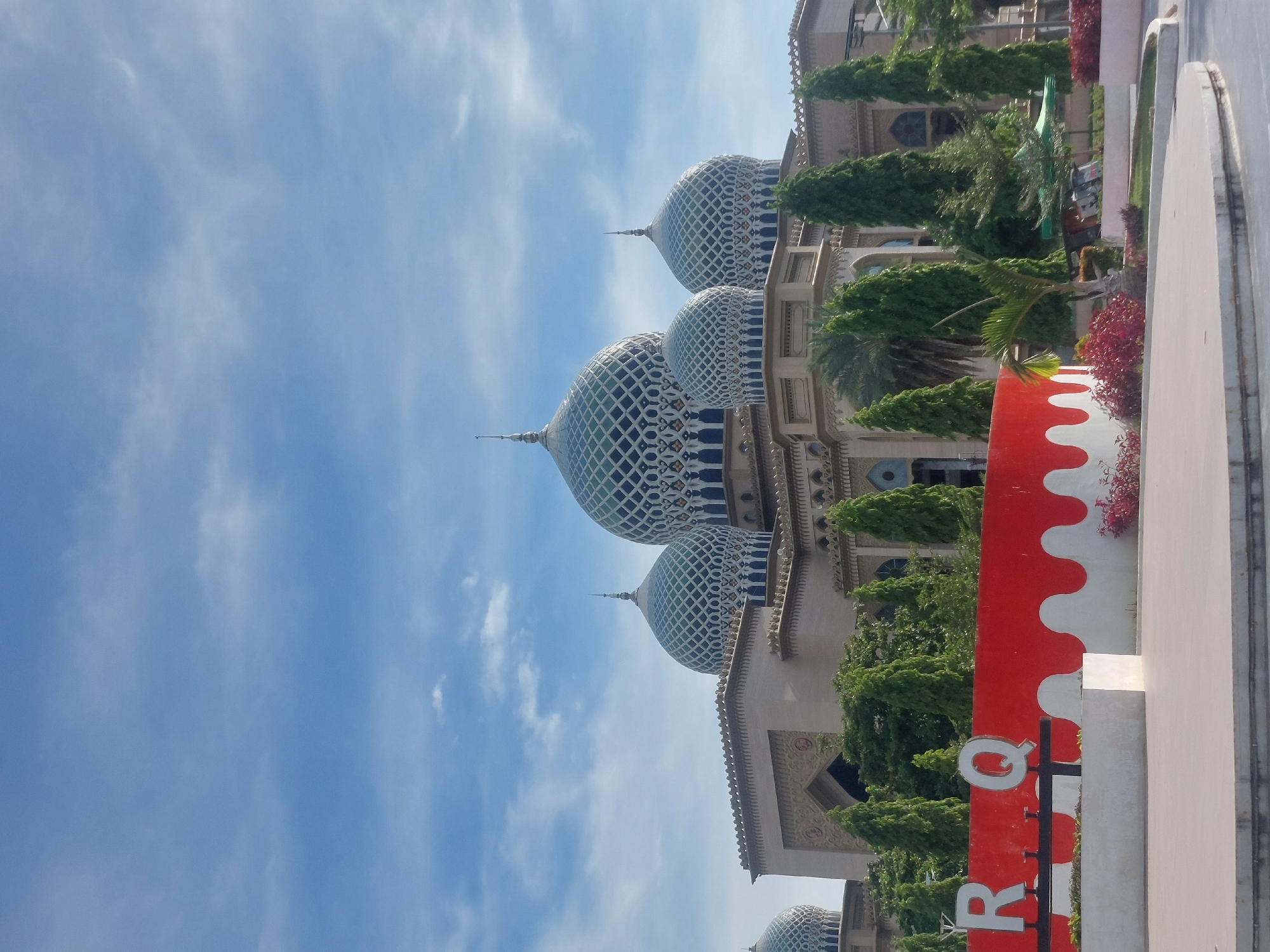 The image depicts the Baiturrahman Grand Mosque in Lhokseumawe, Indonesia, under a partly cloudy blue sky. The mosque features multiple domes with intricate blue and white geometric patterns. In the foreground, a red and white curved wall displays the letters "R" and "Q" in white. Surrounding the mosque are lush green trees, and a paved area is visible in the immediate foreground. The scene is captured during daylight, suggesting an outdoor public space.