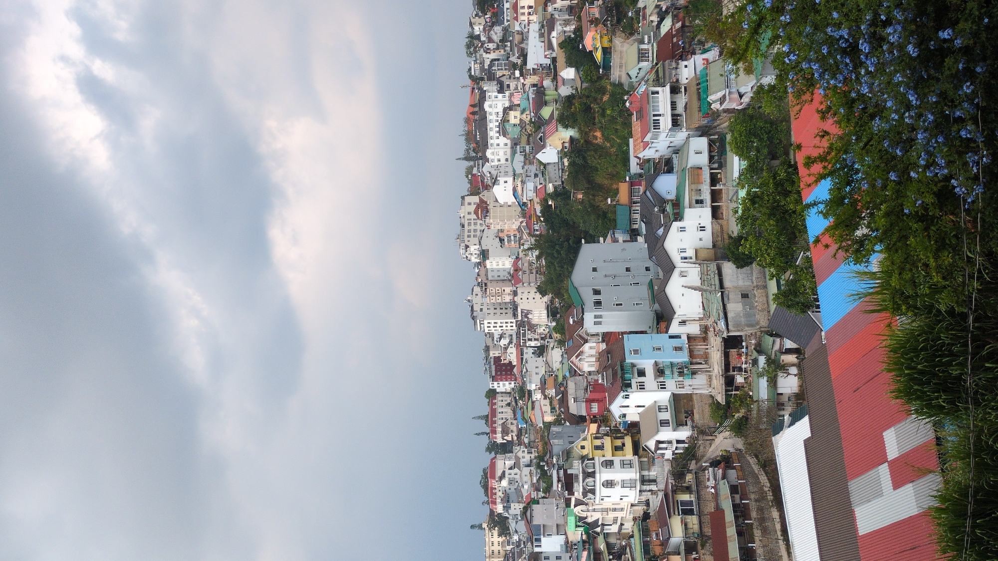 The image depicts a densely packed urban landscape in Da Lat, Vietnam. A mix of residential and commercial buildings with varying heights and colorful roofs are nestled together. Buildings range from small houses to multi-story structures. Trees are interspersed throughout the cityscape. The sky is overcast. A building with red, blue and grey rooftops sits to the bottom right of the frame, surrounded by shrubbery.