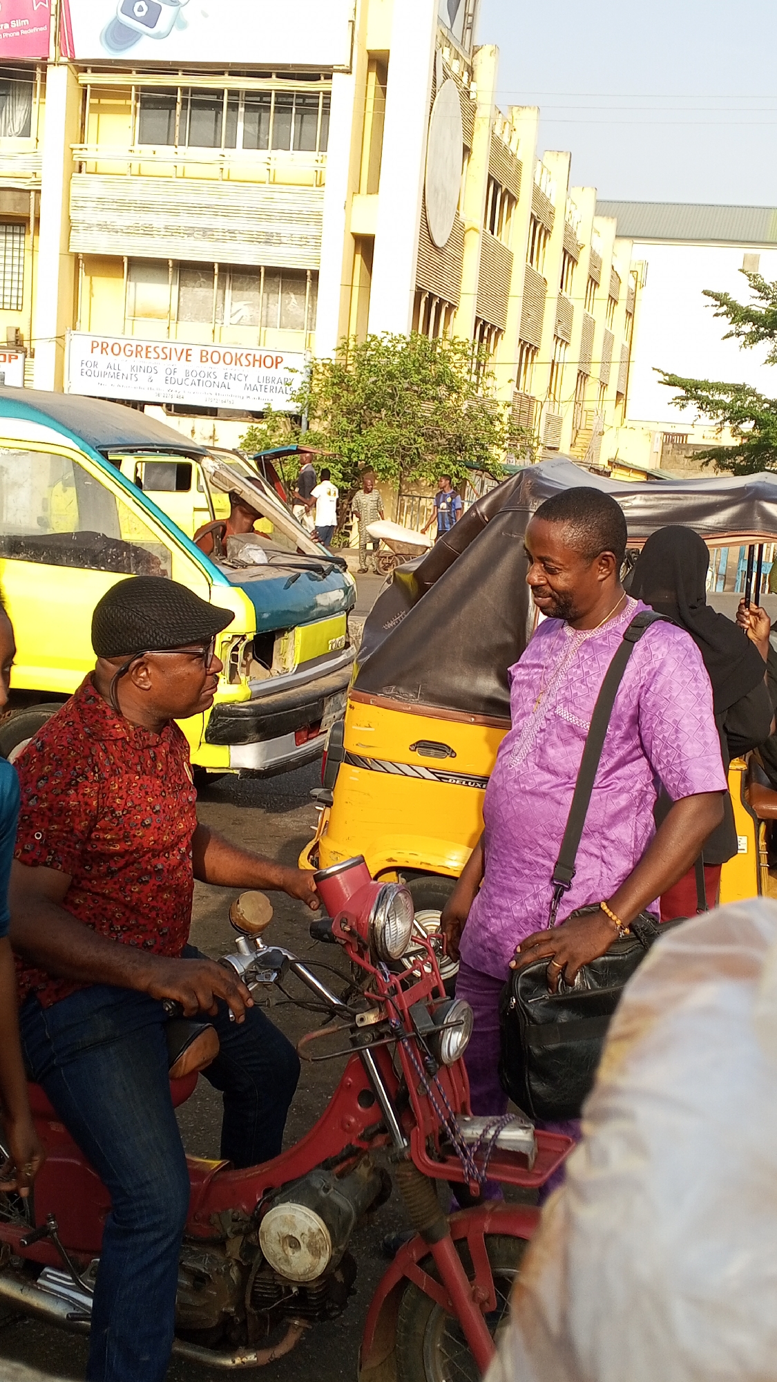 The image, likely taken in the city of Maiduguri, Nigeria, shows a bustling street scene. Two men are engaged in conversation. One is seated on a red motorbike, wearing a red patterned shirt, blue jeans, and a black cap. The other man stands beside the motorbike, dressed in a purple patterned shirt, with a black leather bag slung over his shoulder.
Behind them is a yellow "Keke Napep" or auto-rickshaw with a black hood. Further behind is a yellow and blue minibus. In the background, the "Progressive Bookshop" building is visible, with the text "PROGRESSIVE BOOKSHOP FOR ALL KINDS OF BOOKS ENCY LIBRARY EQUIPMENTS & EDUCATIONAL MATERIALS" displayed. Other people are present in the background, some walking along the street. The environment appears to be a busy commercial area.