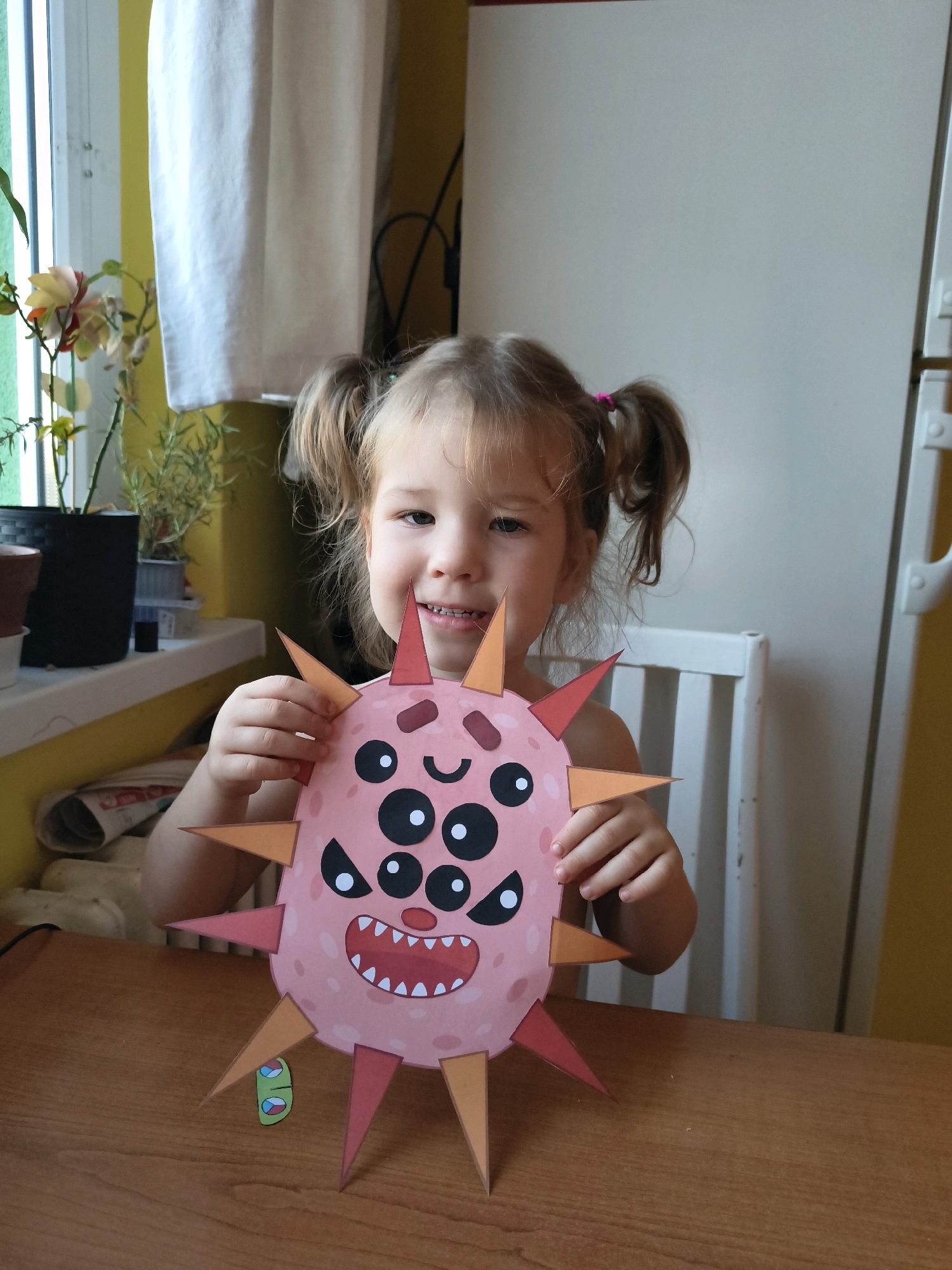 A young girl with blonde hair in pigtails sits at a wooden table, holding a pink, spiky paper craft that resembles a virus or monster. The craft has multiple eyes, a wide mouth with sharp teeth, and several pointed spikes around its perimeter. The girl is smiling at the camera, her teeth visible. She is wearing a dark-colored top, with her shoulders bare. In the background, there is a white chair, a window with a white curtain, and a yellow wall. The overall scene appears to be an indoor setting, likely a home environment. There is no information to suggest the image was taken in Olomouc, Czechia.