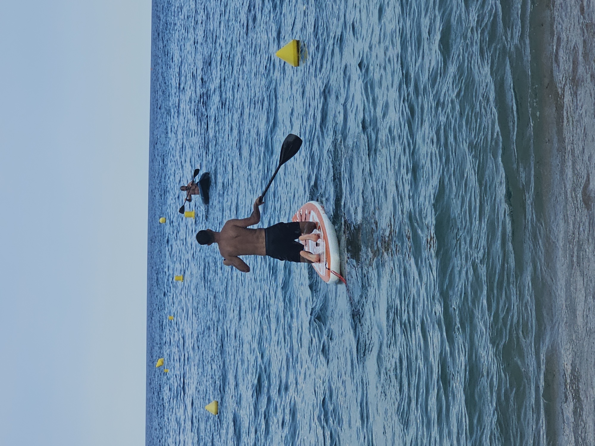 Two men are paddleboarding on the ocean in Blanes, Spain. The man in the foreground is kneeling on a paddleboard, facing away from the camera, holding a paddle. He is wearing black shorts. Another man is in the background, paddling a kayak. Both men are shirtless. There are several yellow buoys in the water. The sky is clear and blue, and the water is choppy. The image appears to have been taken from the shore.