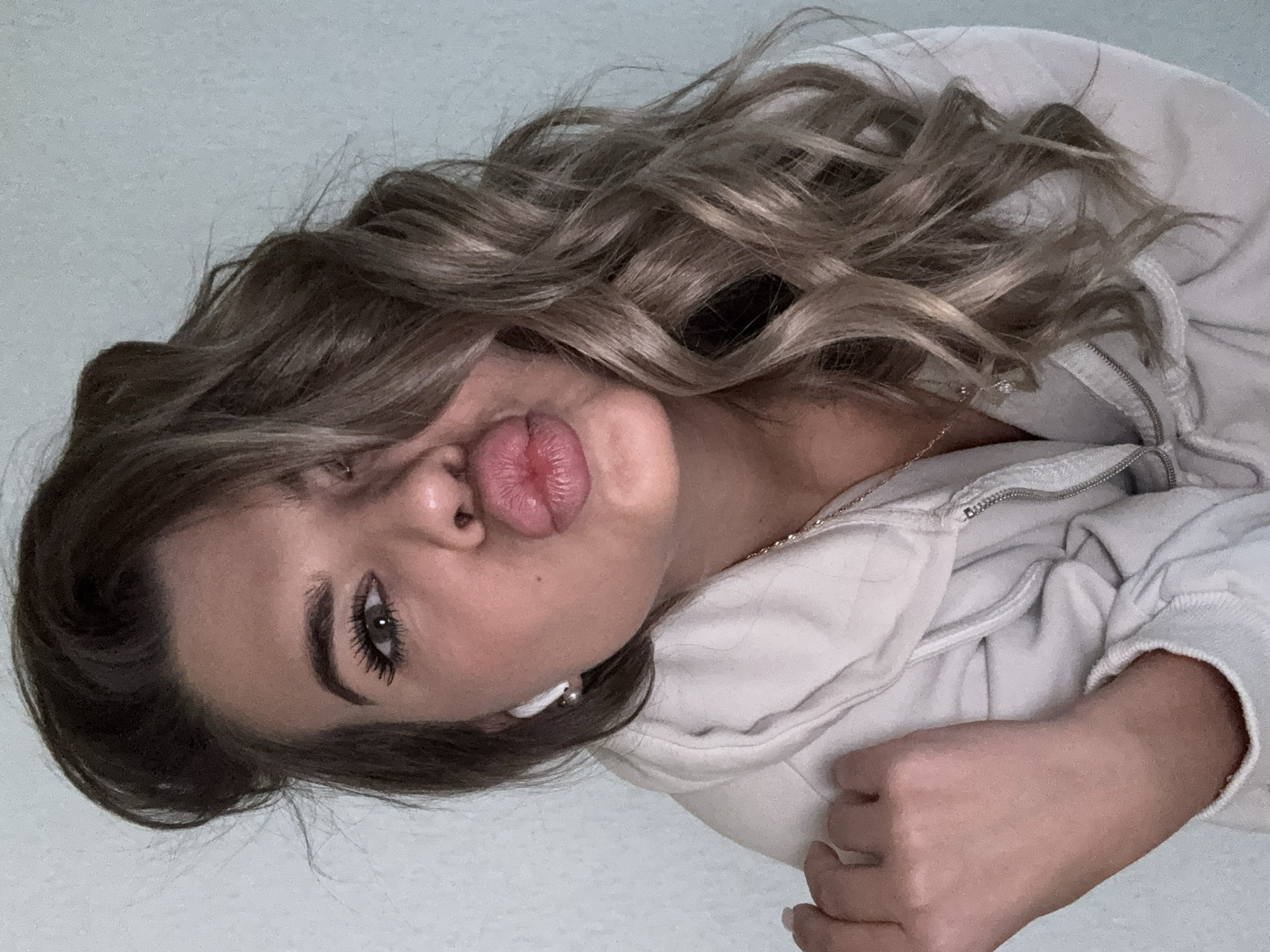 The media file features a close-up, rotated portrait of a young person, presumably taken indoors in Chaikovsky, Russia. The individual, appearing to be female, is centered in the frame, angled with their head towards the left side of the image. They have long, wavy, light brown hair with blonde highlights, which cascades around their face and shoulders.
The person is making a playful "kiss" face, pouting their full, pink lips directly towards the camera. Their left eye, visible and enhanced with dark mascara and eyeliner, looks towards the viewer. Dark, well-defined eyebrows frame the eye. The person has clear skin.
They are wearing a light-colored (likely white or light grey) zippered hoodie, with the zipper partially open. A thin, silver-colored chain necklace is visible against their skin. A white earbud is seen in their right ear. In the lower right corner of the image, a hand with light-colored, manicured nails is visible, suggesting the person might be holding the camera or posing casually. The background is a plain, light-colored wall, providing a simple, uncluttered setting for the portrait. The overall lighting is bright and even, indicating either natural daylight indoors or good artificial illumination. No visible text is present in the image. The pose and expression convey a casual and cheerful mood.