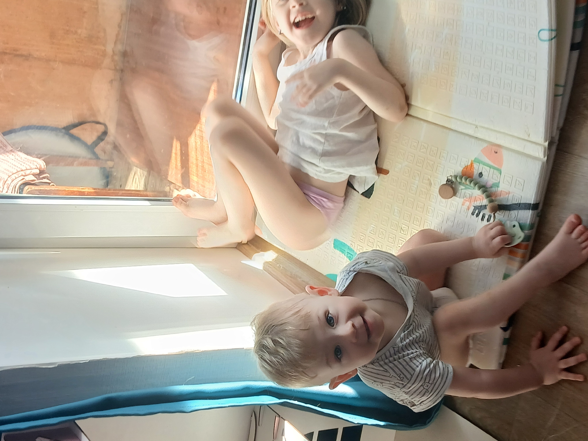 Two young children are in a room with a large window. One child, a girl, is lying on a play mat. She wears a white tank top and light purple underpants. She is smiling and appears to be playing. The second child, a baby, is sitting on a blue padded surface. The baby wears a striped shirt. The baby is also smiling and appears to be playing. Sunlight streams in through the window, suggesting daytime. A bag is visible near the window. The location is assumed to be in Chaikovsky, Russia, based on the image title.