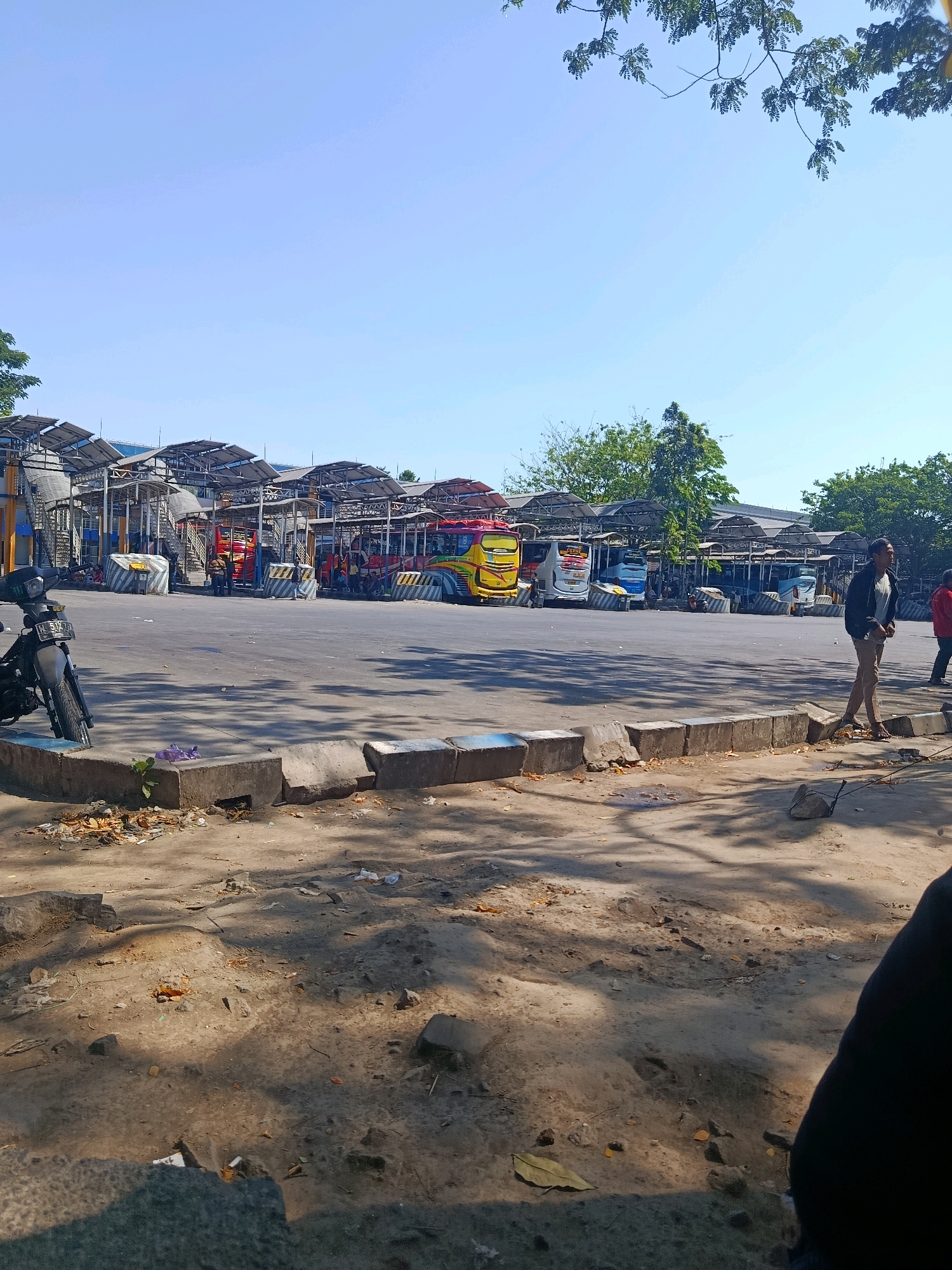 The image depicts an outdoor scene at a bus terminal in Bungurasih, Indonesia. The foreground shows a rough, sandy surface with scattered debris, including leaves, plastic bags, and stones. A concrete barrier, some sections of which are broken, runs horizontally across the mid-ground, with blue painted markings. To the left, a parked motorcycle is visible.
In the background, a row of covered bus bays stretches across the frame. Several buses are parked under these shelters, including a yellow and red bus with "PO Rosalia Indah" branding and a white and blue bus. People are seen standing near the buses and some are seated. The bus terminal is characterized by its metal structure with corrugated roofs, designed to provide shade. The environment appears to be a sunny day, with clear blue skies overhead and shadows cast on the ground from trees and structures. The overall impression is that of a busy transportation hub, albeit one with visible signs of wear and tear.