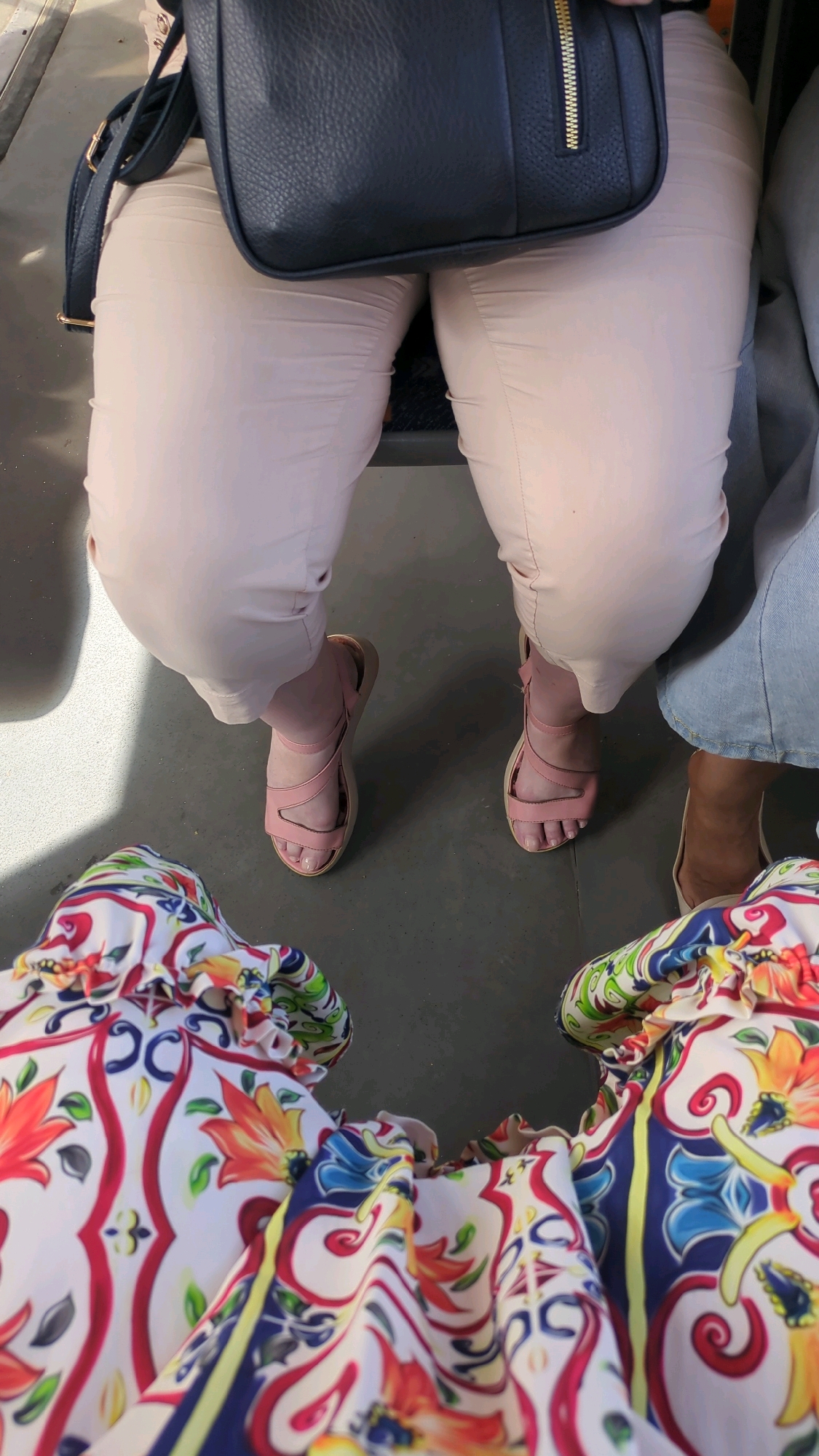 The image shows a close-up, eye-level perspective of two individuals from the waist down, seated in what appears to be public transportation. The person in the foreground is wearing a vibrant, multi-colored patterned top with a ruffled neckline. The person behind them is wearing light-colored pants and pink strappy sandals. A dark blue purse with a gold zipper is visible resting on the lap of the person in the light-colored pants. The ground beneath them is a dark, textured surface. The lighting suggests an indoor environment, possibly a bus or train carriage, with natural light filtering in from an unseen source, casting shadows. No specific landmarks or identifying features place this scene within Chișinău, Moldova; the environment is generic for urban public transport.