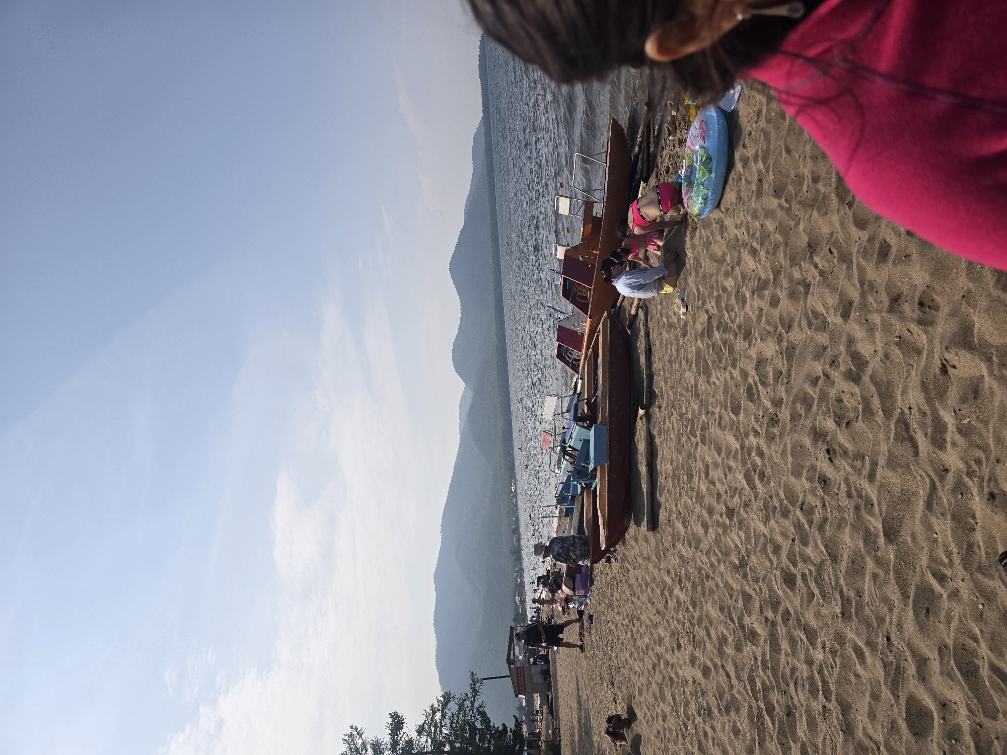 The image depicts a sunny day at a sandy beach on a lake in Maksimiha, Russia. The scene is viewed from a slightly elevated perspective, looking out over the water towards a mountainous horizon. The water is calm, with gentle ripples on the surface. Several pedal boats are lined up on the shore, some with people sitting in or near them.
In the foreground, the sand is rippled and textured, indicating a natural beach environment. A person wearing a bright pink top is visible on the right side of the frame, partially obscuring the view. Several other individuals, including children and adults, are scattered along the shoreline. Two young children are near a pedal boat, seemingly playing in the sand. One child wears a pink swimsuit, and another wears a blue shirt and yellow shoes. An inflatable ring with a floral pattern is also present on the sand. Further down the beach, more people are visible, some standing and some sitting. There are trees on the far left side of the image and some structures suggesting a beachside establishment.
The time of day appears to be late afternoon or early evening, given the angle of the sunlight casting long shadows and the warm glow on the sand. The sky is clear and light blue, with a few wispy clouds. The overall mood of the scene is relaxed and leisurely, suggesting a day spent enjoying outdoor recreation by the lake. No discernible text is visible in the image.
