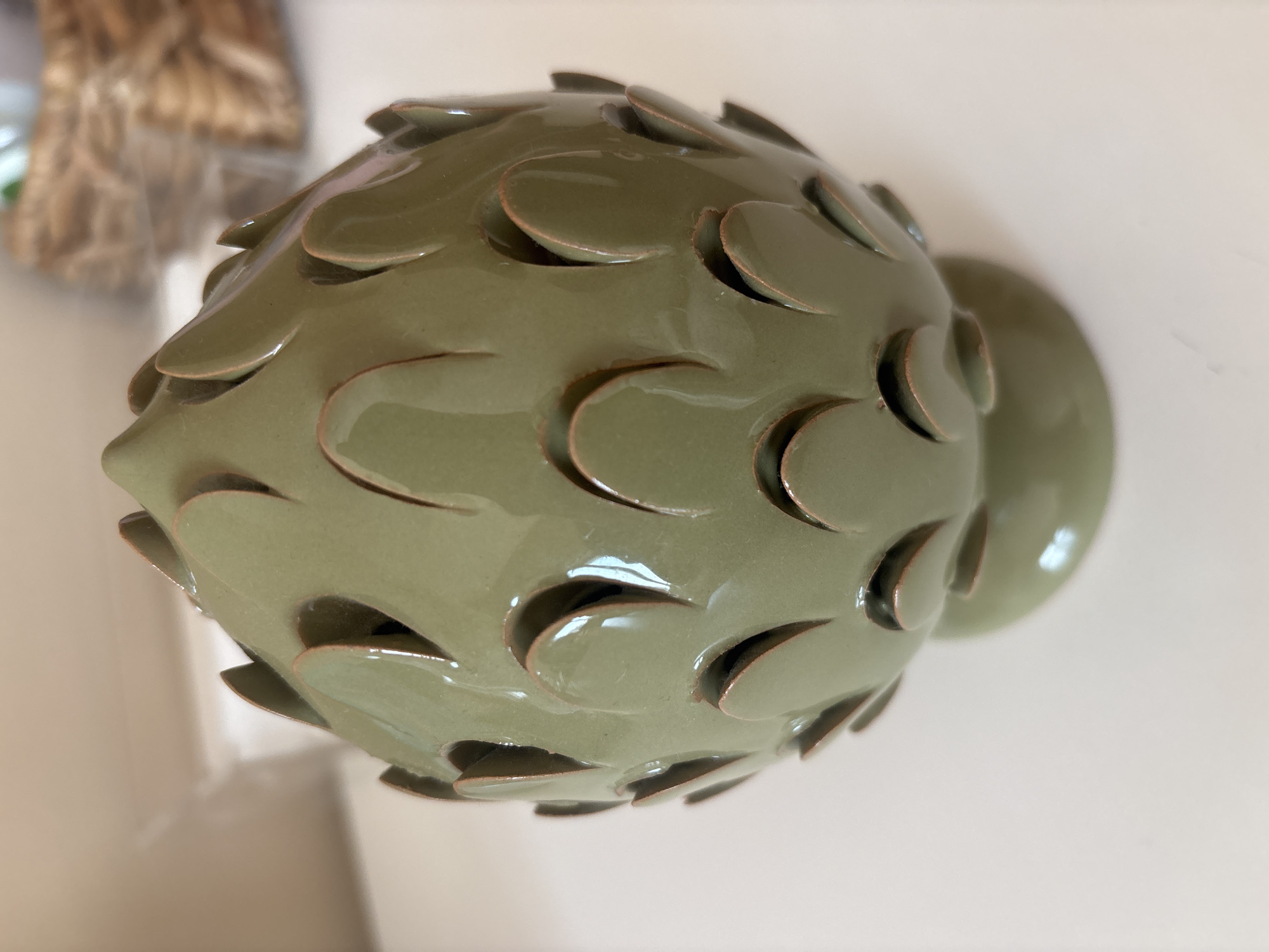 A close-up, eye-level studio shot features a decorative ceramic artichoke object. The object is a light olive green with a glossy finish and is shaped like an artichoke, complete with overlapping leaf-like indentations that create openings. The bottom of the object flares slightly into a small pedestal base. The object sits on a white surface, likely a table. The background is out of focus, showing a hint of a light-colored wall and a blurred wicker basket on the right. The image was taken indoors and does not provide any information about its location in Dubai, United Arab Emirates, or the presence of any people.