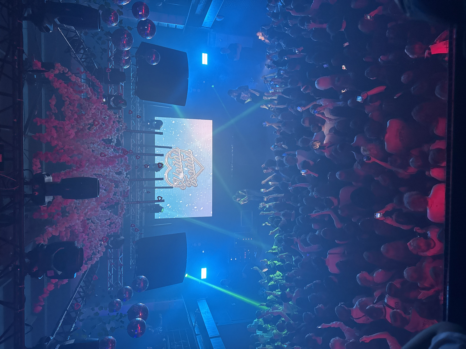 The image depicts a vibrant indoor concert or club event, captured from an elevated perspective looking down upon the stage and a large, engaged audience. The scene is dark, illuminated by dynamic stage lighting in blue, green, and red, along with laser beams cutting through the air.
The stage area, positioned on the left, features a prominent large LED screen displaying the stylized text "Lovely Sunday," which appears to include a heart graphic. To the left of the screen, a significant volume of pink or reddish decorative elements, resembling either confetti or artificial flower petals, is suspended in the air, creating a festive visual effect. Above the stage, large speakers, various stage lighting fixtures, and multiple reflective disco balls are visible, attached to a metal truss system.
A dense crowd of people fills the right side of the frame, extending towards the stage. Most attendees are seen from behind, with many holding up their smartphones to record or photograph the event. The crowd is actively engaged, with some individuals raising their hands. The red stage lighting casts a warm glow over sections of the audience.
The event is taking place at night, typical for such performances. The atmosphere is energetic and lively. This scene unfolds in Vladivostok, Russia.