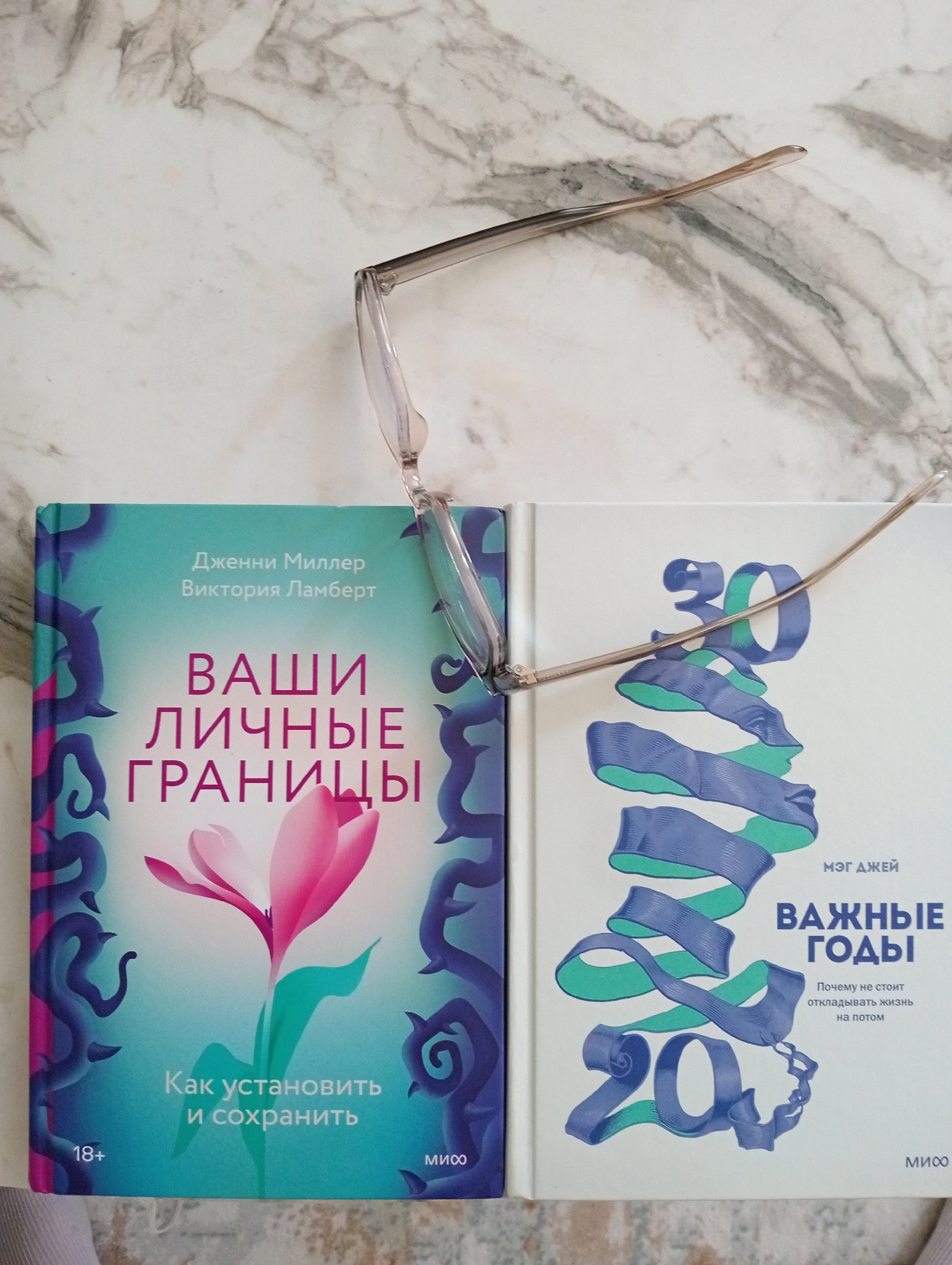 The image shows two books on a marble surface, with a pair of eyeglasses resting on top of them. The book on the left has a teal cover with a pink and white flower and the title "ВАШИ ЛИЧНЫЕ ГРАНИЦЫ" (Your Personal Boundaries) written in large pink font. The book on the right has a white cover with a blue and green ribbon design and the title "ВАЖНЫЕ ГОДЫ" (Important Years) in large blue font. The eyeglasses are clear-rimmed and are positioned diagonally across the books. The background is a marbled surface, typical of countertops or tabletops. The scene is a close-up shot, with no indications of a specific location within Silifke, Turkey or any other city. There are no people or actions depicted in the image.